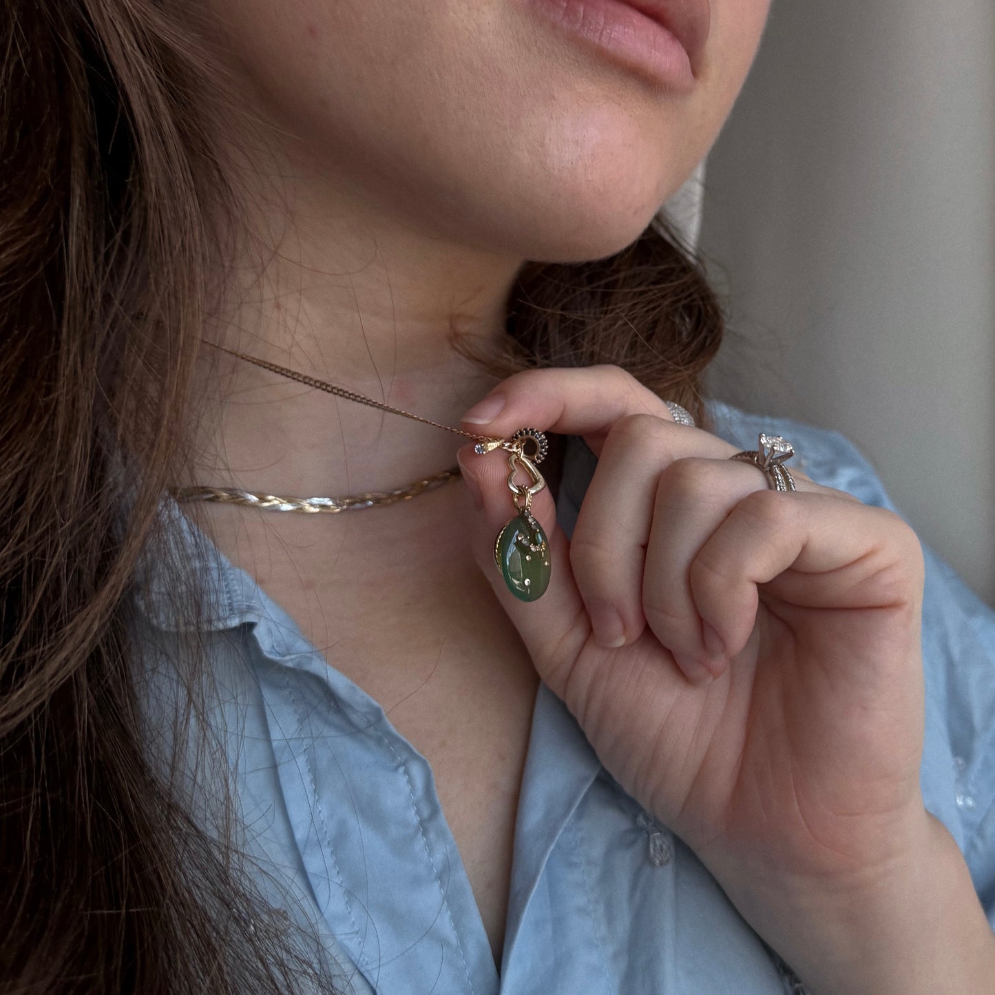 A person in a blue shirt holds up a necklace with a green pendant, wearing the Charlie and Marcelle 14k Littlest Birthstone Ring Charm and a ring with a large clear stone. Their brown hair falls over their shoulder.