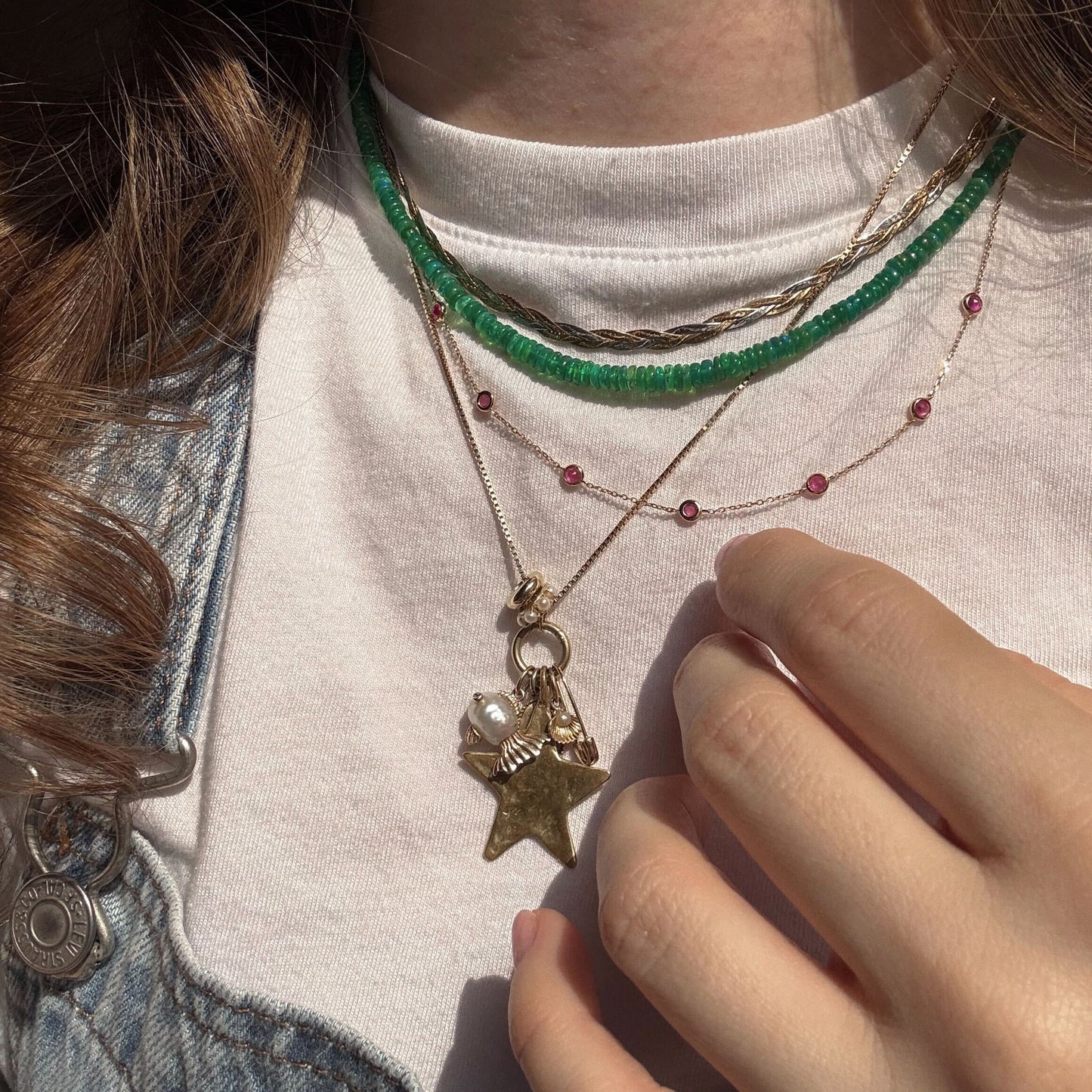 A person wears layered necklaces: a green beaded choker, a delicate 9k solid yellow gold chain with red beads (Solid Gold Birthstone Station Necklace Sample), and a gemstone charm necklace. Theyre in a white t-shirt and denim jacket, touching the charms.