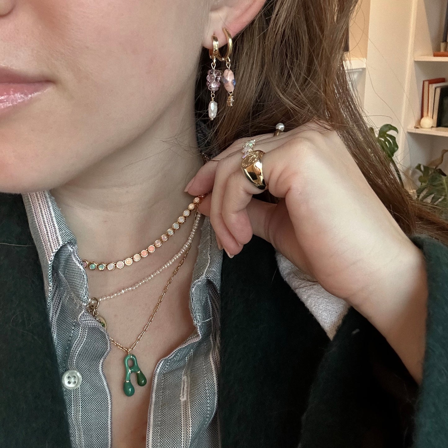 A woman wears layered necklaces, including the Bubble Letter Necklace, a beaded choker, and a green pendant. She also sports pearl and gem earrings and multiple rings, posing with her hand near her face in a green coat over a button-down shirt.