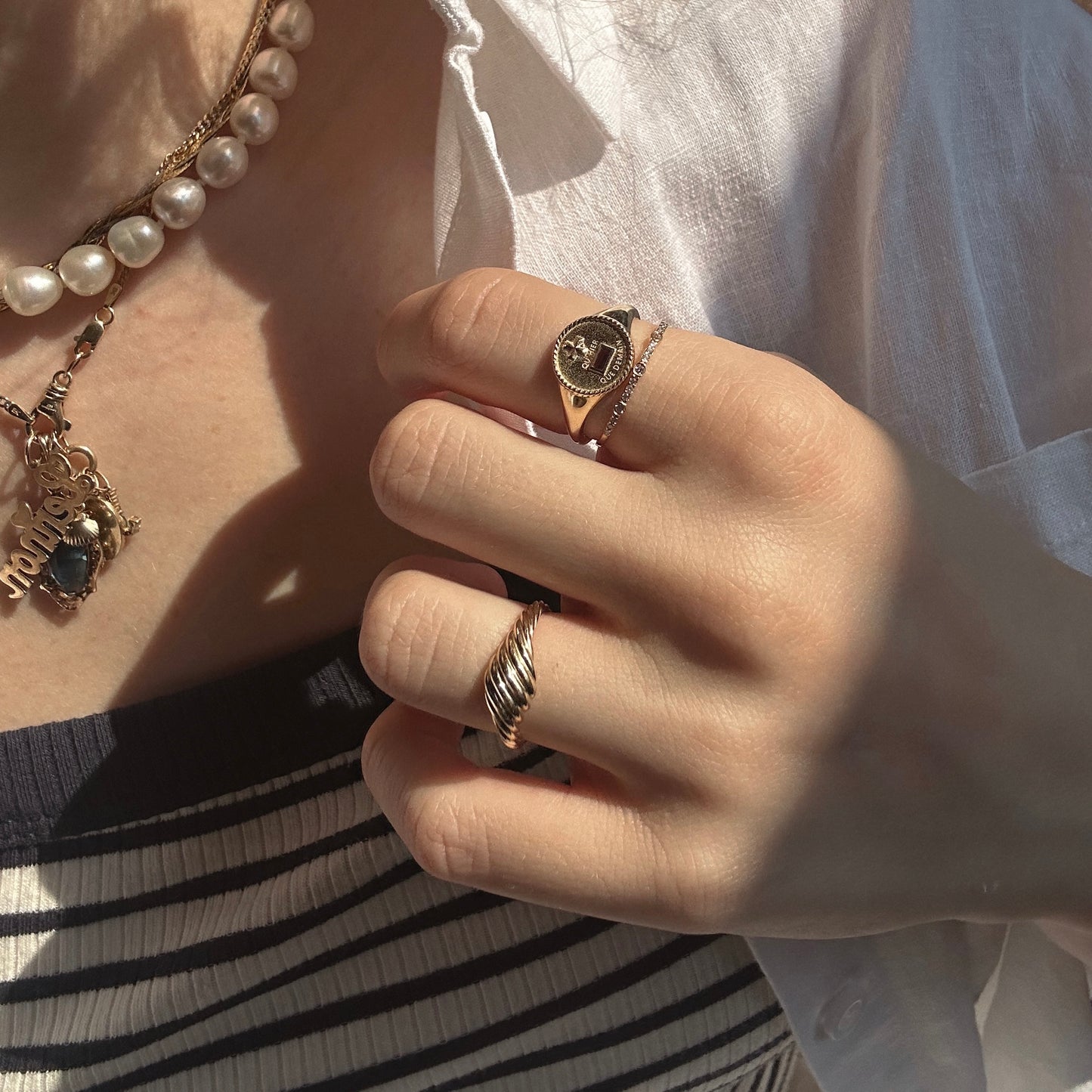 A hand with a Solid Gold Delicate White Sapphire Stacking Band and a pearl necklace, wearing a white shirt over a black and white striped top, is shown in sunlight. The jewelry also includes a pendant with a dark stone and name charm.