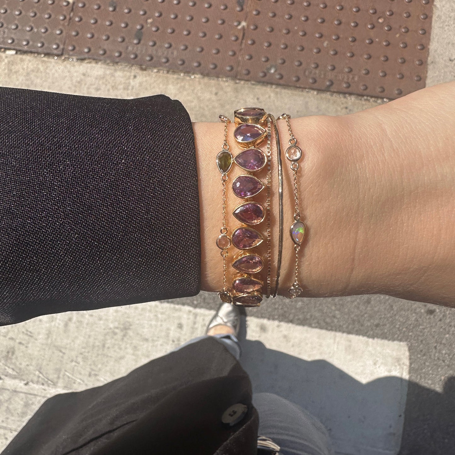 A person in a black sleeve wears three bracelets, including the Solid Gold Structured Bracelet, a leather band with purple teardrop gems, and a delicate chain, while standing on a city sidewalk near a textured metal plate.