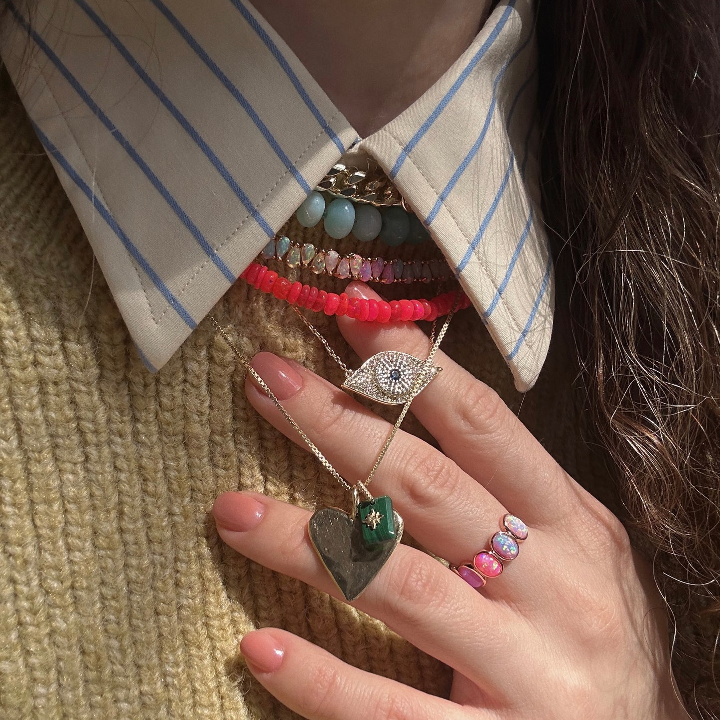 A close-up of a person in a beige and blue striped collared shirt and mustard knit sweater, layered necklaces with colorful beads and an eye pendant, plus rings including the Rose Gold Opal Ombre Candy Band with vibrant stones.