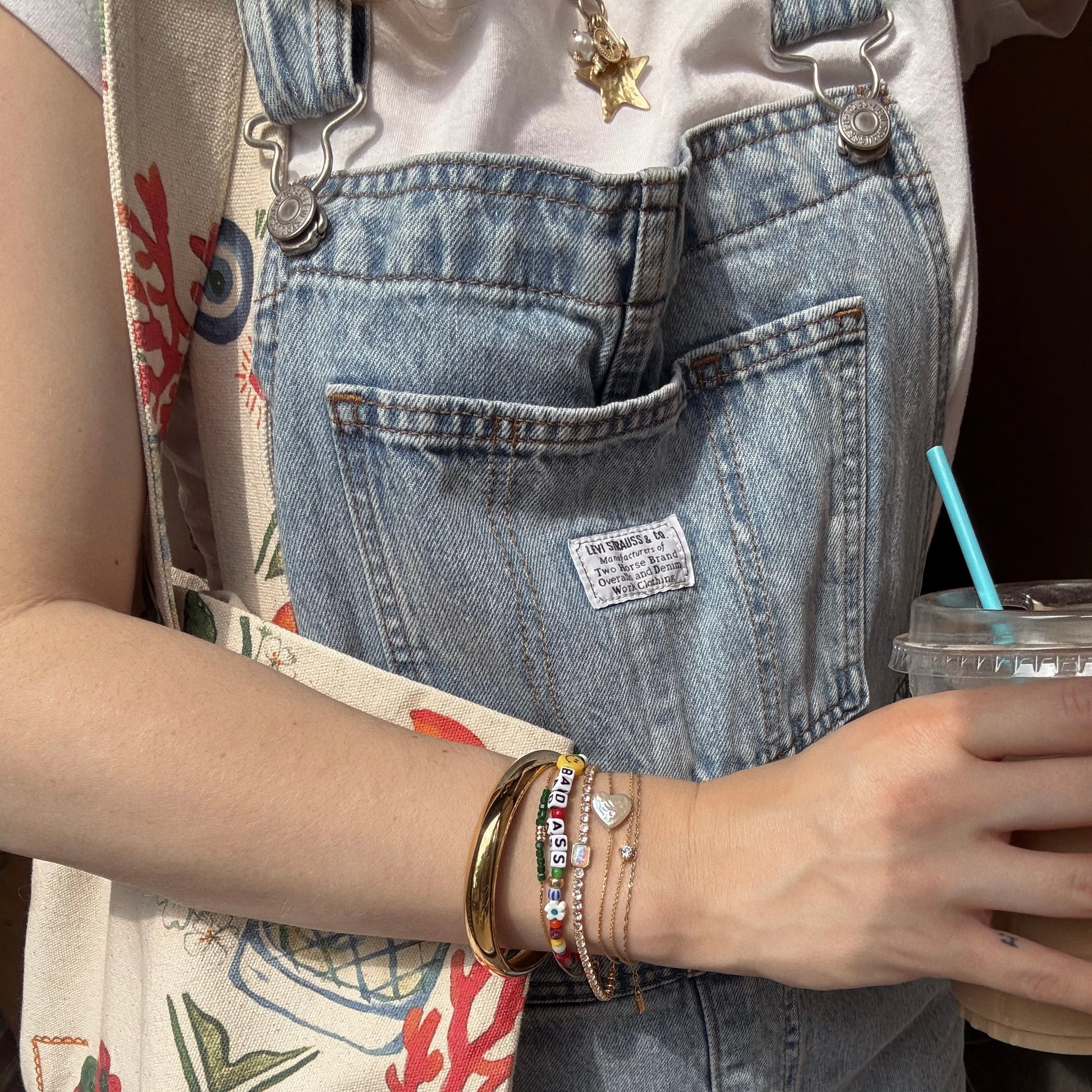 A person in denim overalls and a white T-shirt holds a drink with a blue straw. They wear the Handmade Beaded Bada$$ Bracelet, a 14k gold plated star necklace, and carry a red and green patterned tote bag.