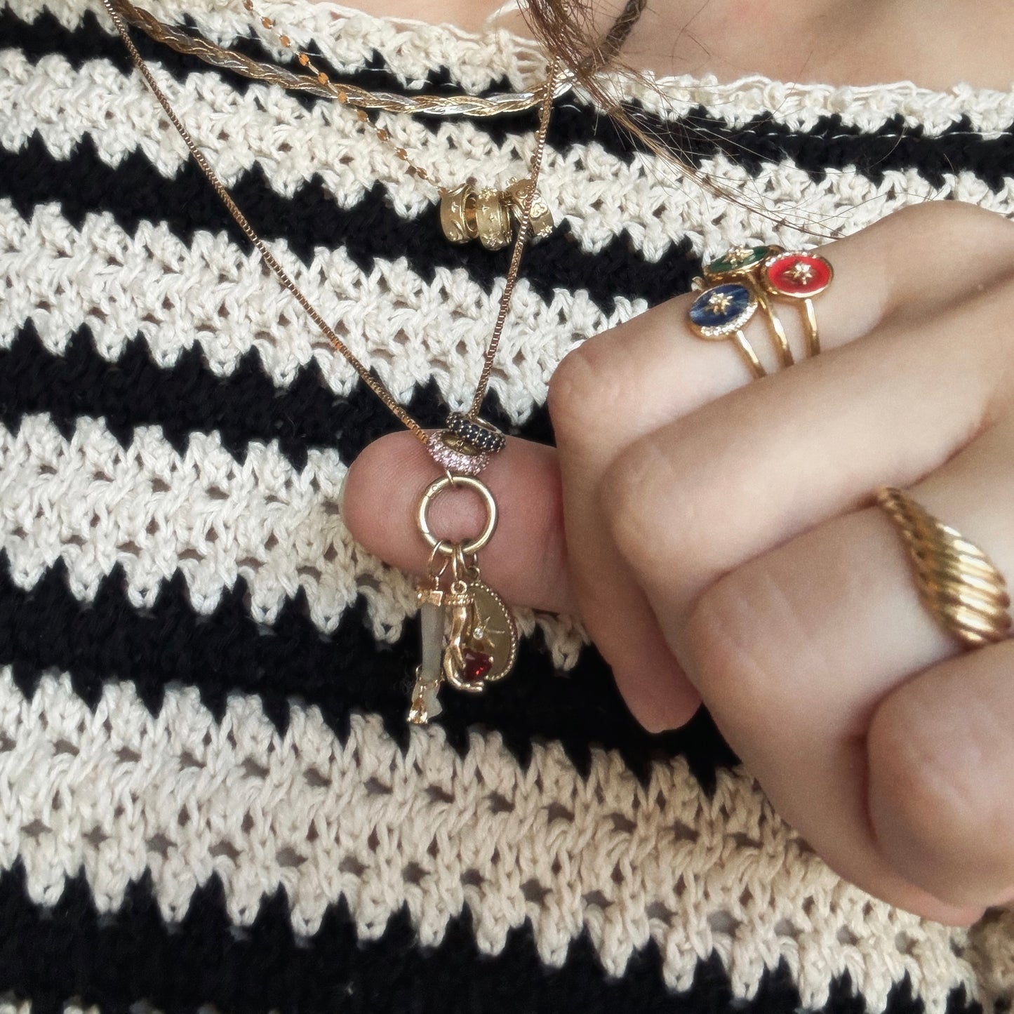 A person wearing a black and white striped knit top holds the Solid Gold Body Language Charm necklace, exclusive to Local Eclectic, with multiple gold charms. They also wear gold rings, one with red, blue, and white floral designs.