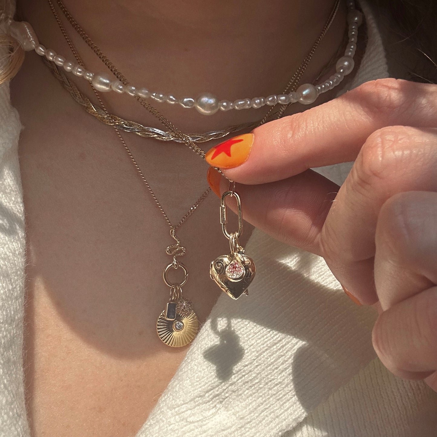 A person wears layered gold and pearl necklaces, featuring the Solid Gold Charm Clip personalized necklace, as they hold a heart-shaped gemstone locket. Their orange nail with a red star stands out against cream-colored fabric.