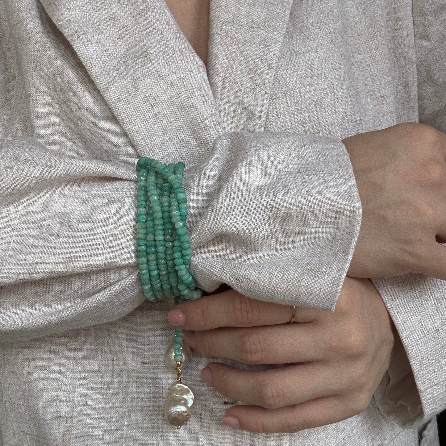 A person in a light beige textured blazer wears the Gemstone Tie Necklace, featuring multiple green bead strands and a baroque pearl pendant charm on their wrist.