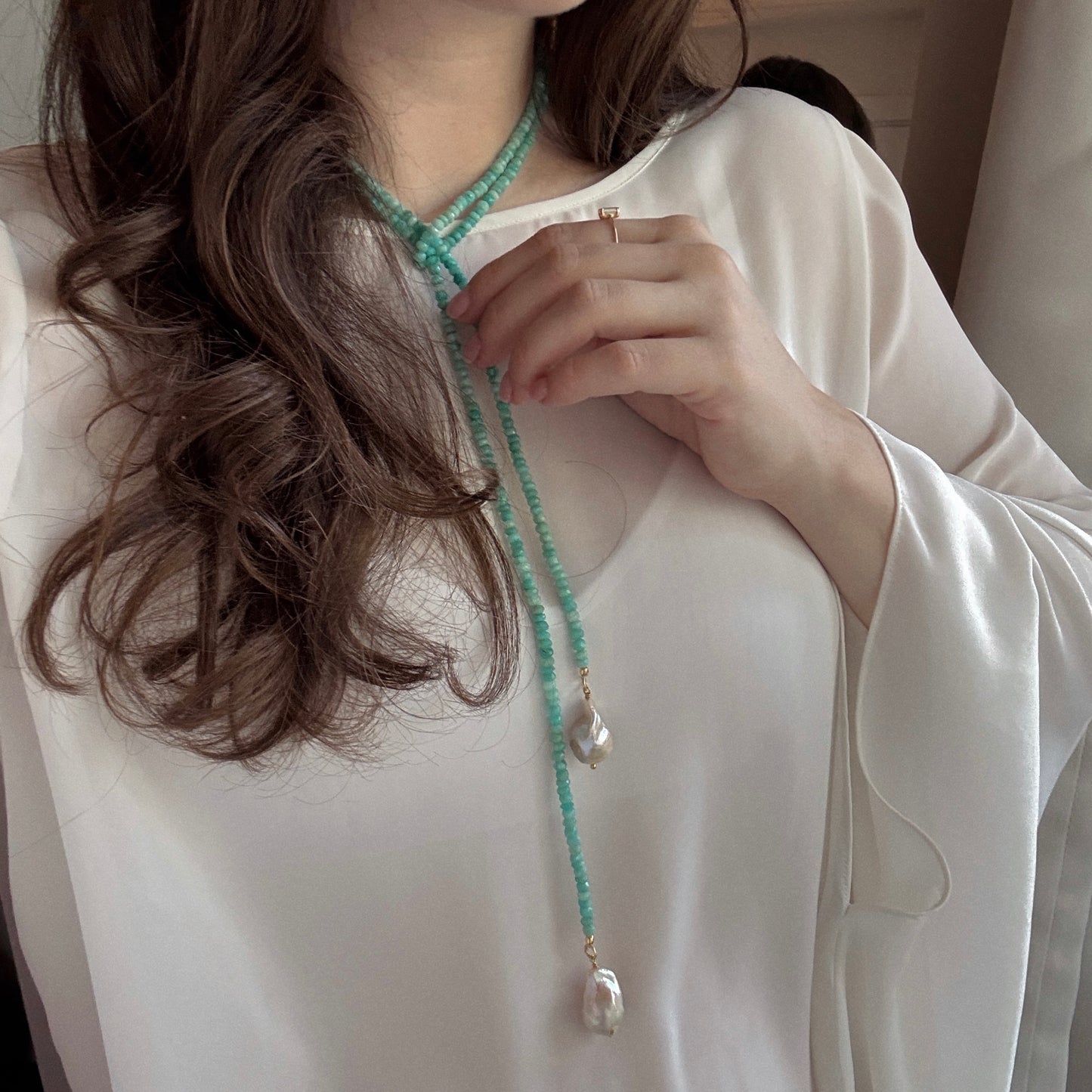 A woman with wavy brown hair draped over her shoulder wears a flowing white blouse and holds the Gemstone Tie Necklace, featuring pearl pendants. She also wears a simple gold ring on her finger.