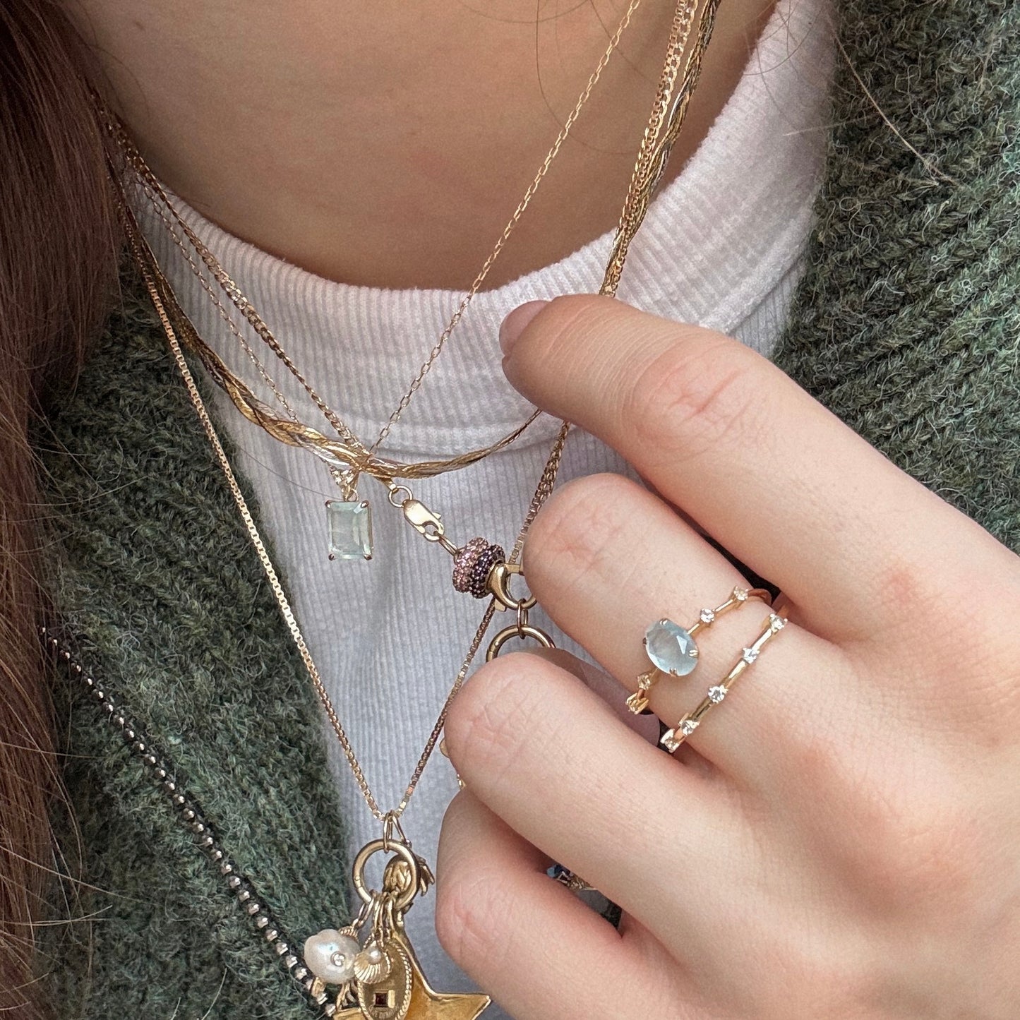 A person wears layered gold necklaces with charms and the Solid Gold 2025 March Capsule Classic Aquamarine Statement Ring among several delicate gemstone rings, paired with a white ribbed top and green sweater.