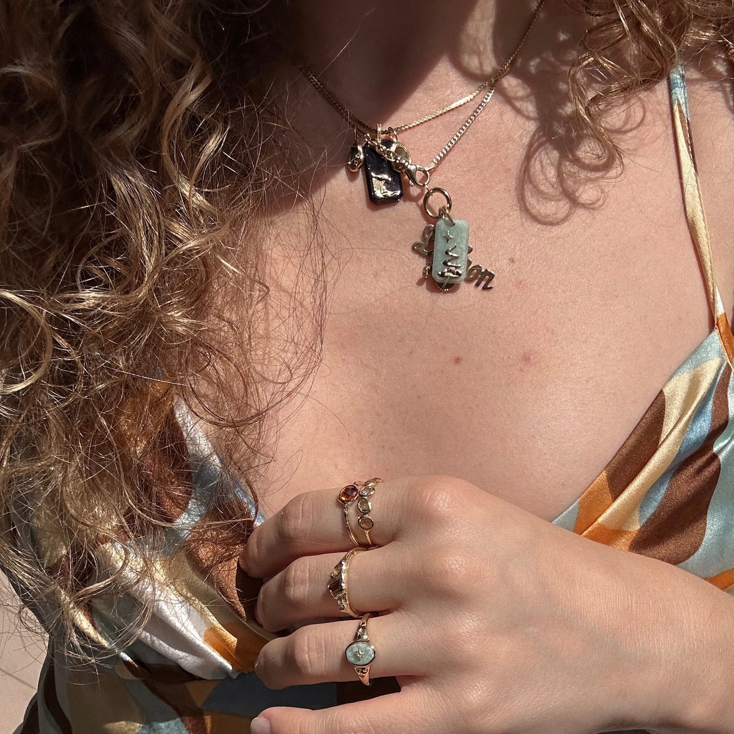 A person with curly hair wears layered gold necklaces, including the Solid Gold Crystal Element Charm, along with rings and a patterned dress. Their hand rests on their chest, drawing attention to the jewelry and vibrant fabric.