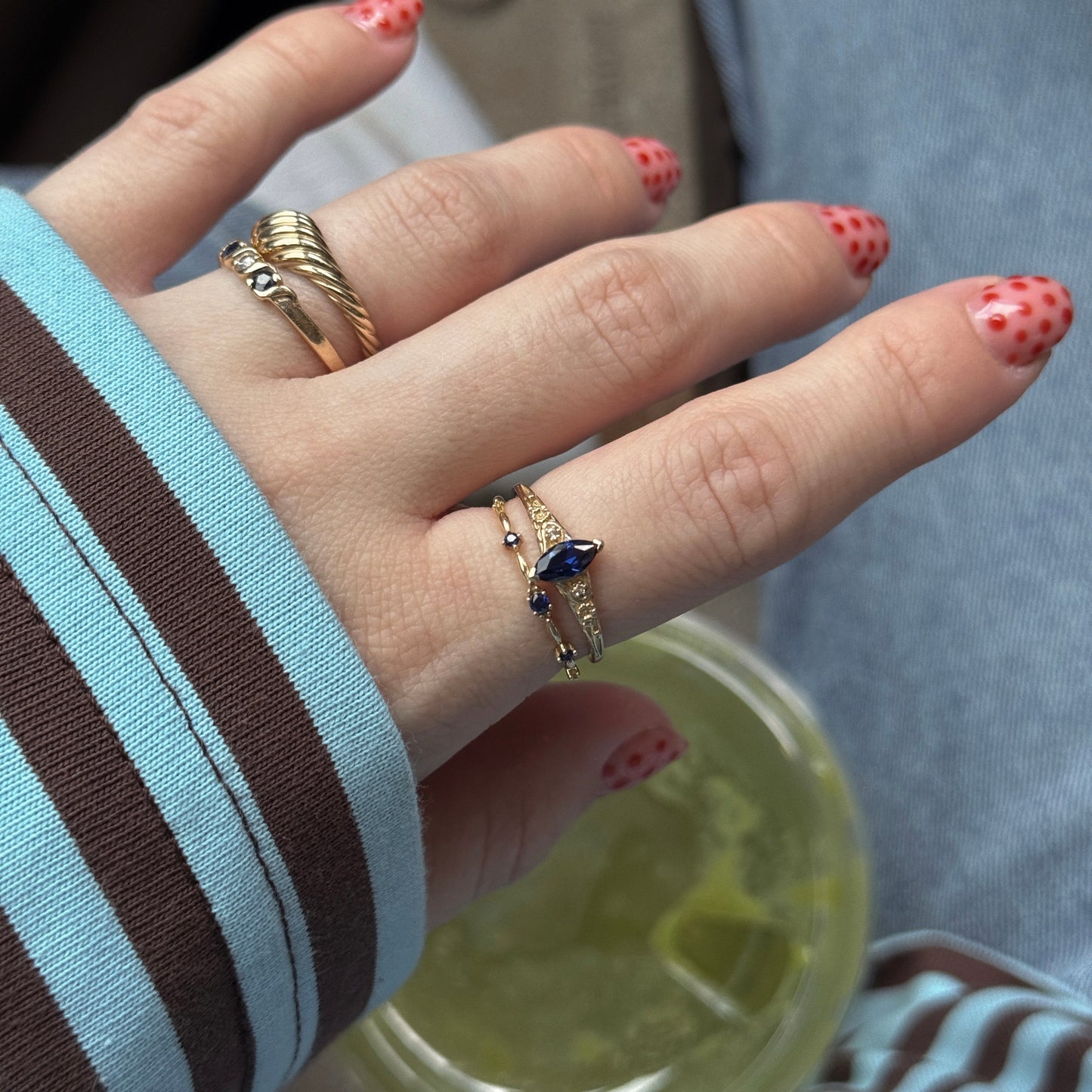 A hand with pink, red-dotted nails wears the Solid Gold 2025 September Capsule Classic Blue Sapphire Statement Ring and two other gold rings, resting on a striped sleeve while holding a drink with lime ice cubes; blue jeans appear in the background.