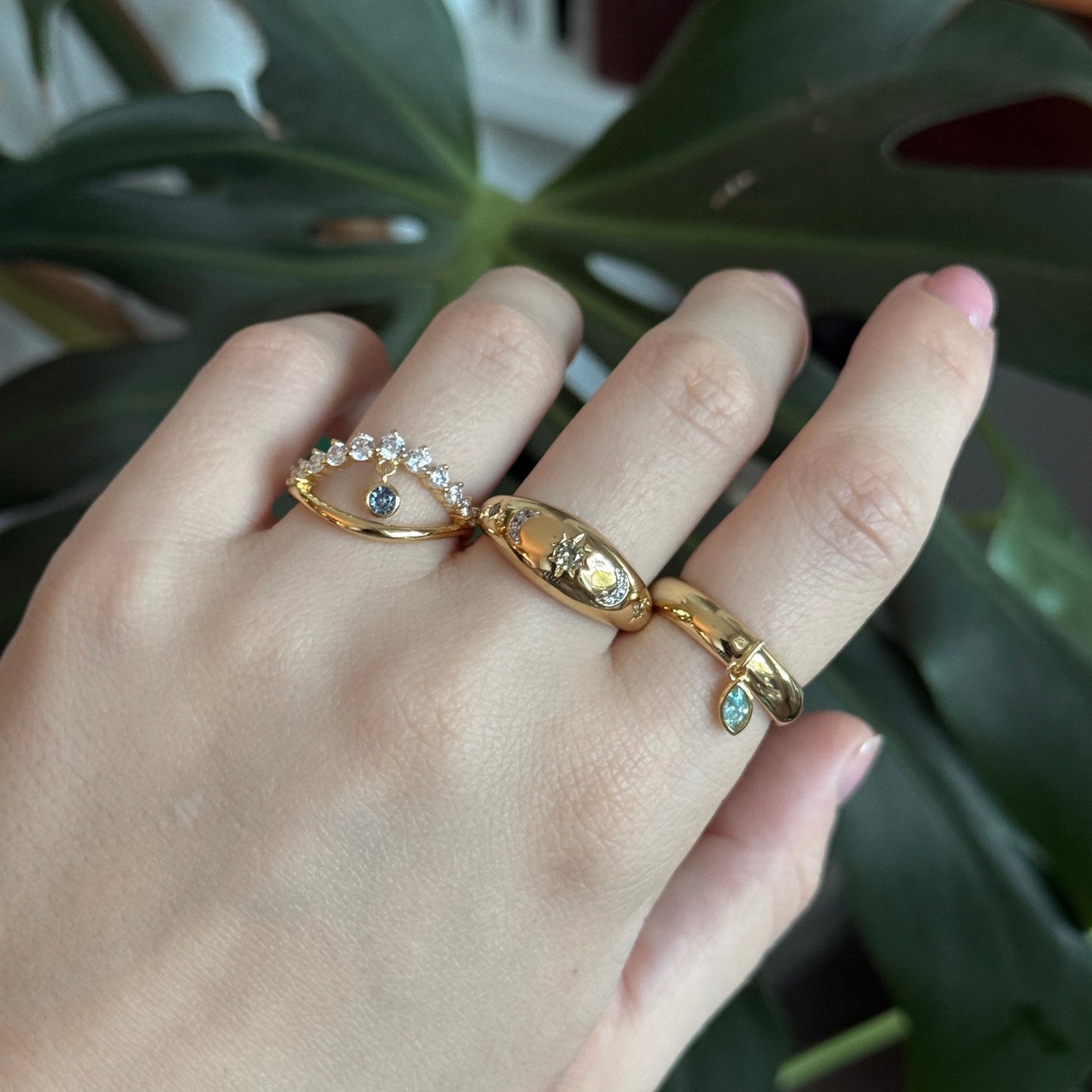 A hand with manicured nails displays the Moonstruck Bold Dome Ring, shining with crystals and colored stones, amid lush green monstera leaves.