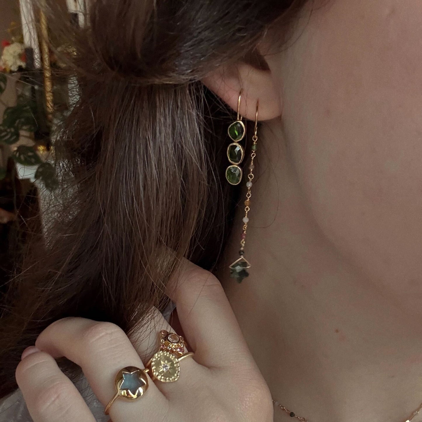 A close-up of a person’s ear and hand wearing Amber Earrings—yellow gold plated, with green tourmaline stones and a star-shaped charm—plus gold rings, including one with a star-shaped stone, against a softly blurred background.