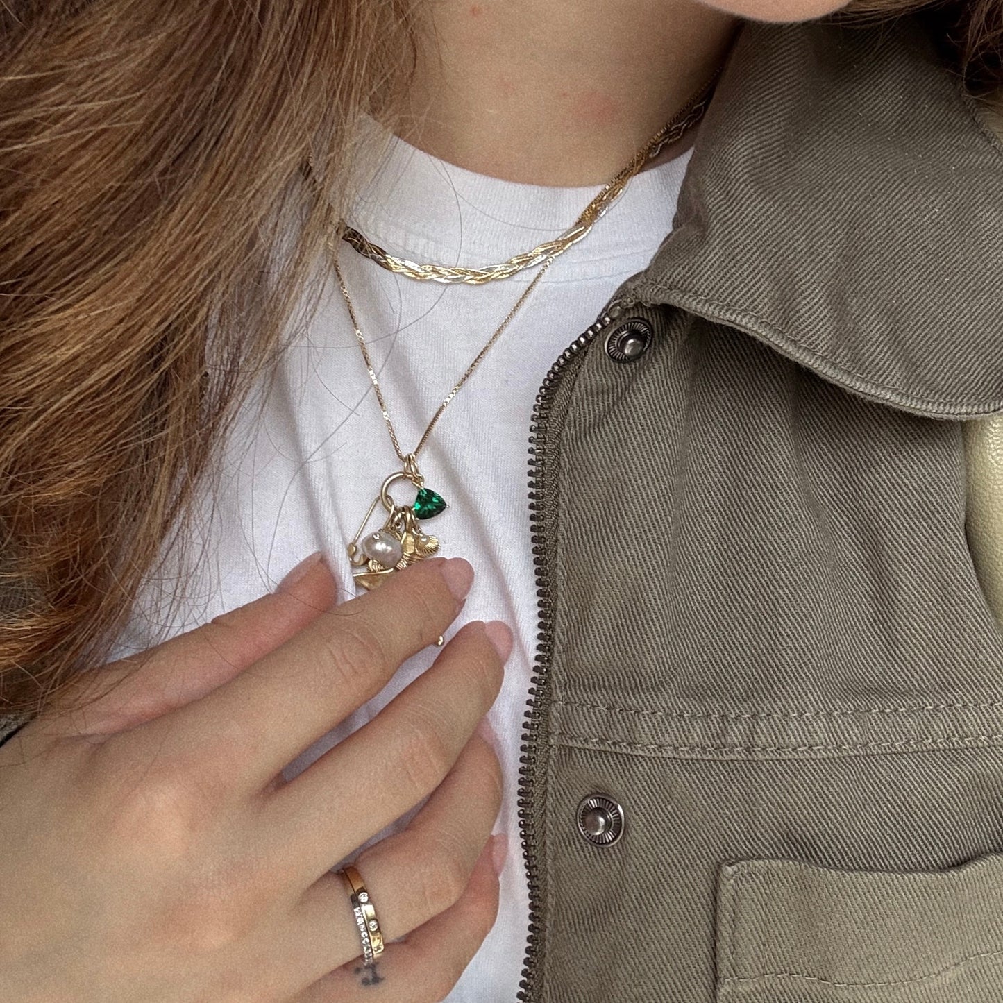 A close-up of a person wearing layered gold necklaces, including the Family Gold Solid Gold Trillion Birthstone Charm. They have long brown hair, a white shirt, olive green jacket, and hold a pendant with a silver ring on their finger.