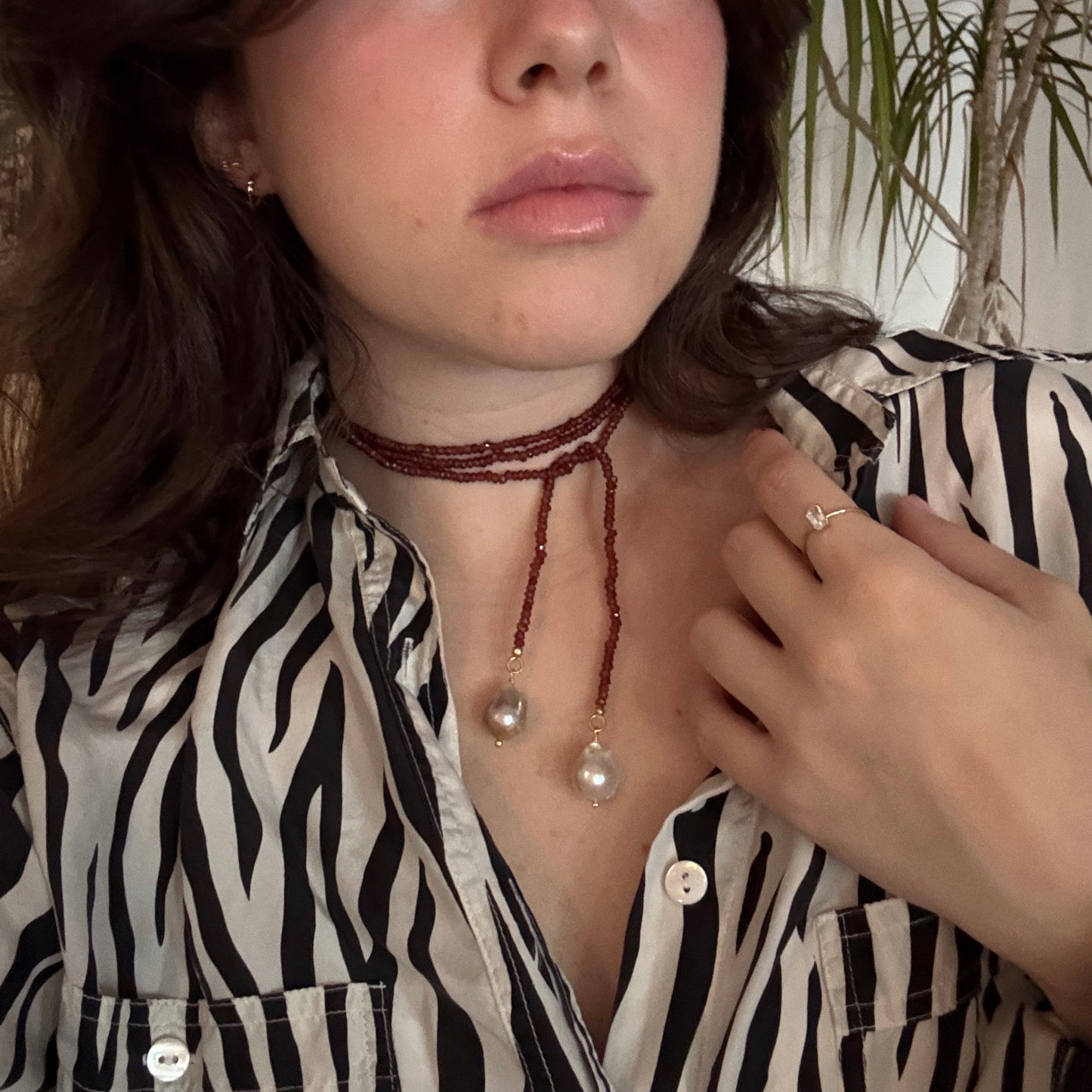 Wearing the Gemstone Tie Necklace, a person pairs it with a zebra-print shirt. Their hand, showcasing a ring, is poised near their neck, while a tall plant creates an interesting backdrop.