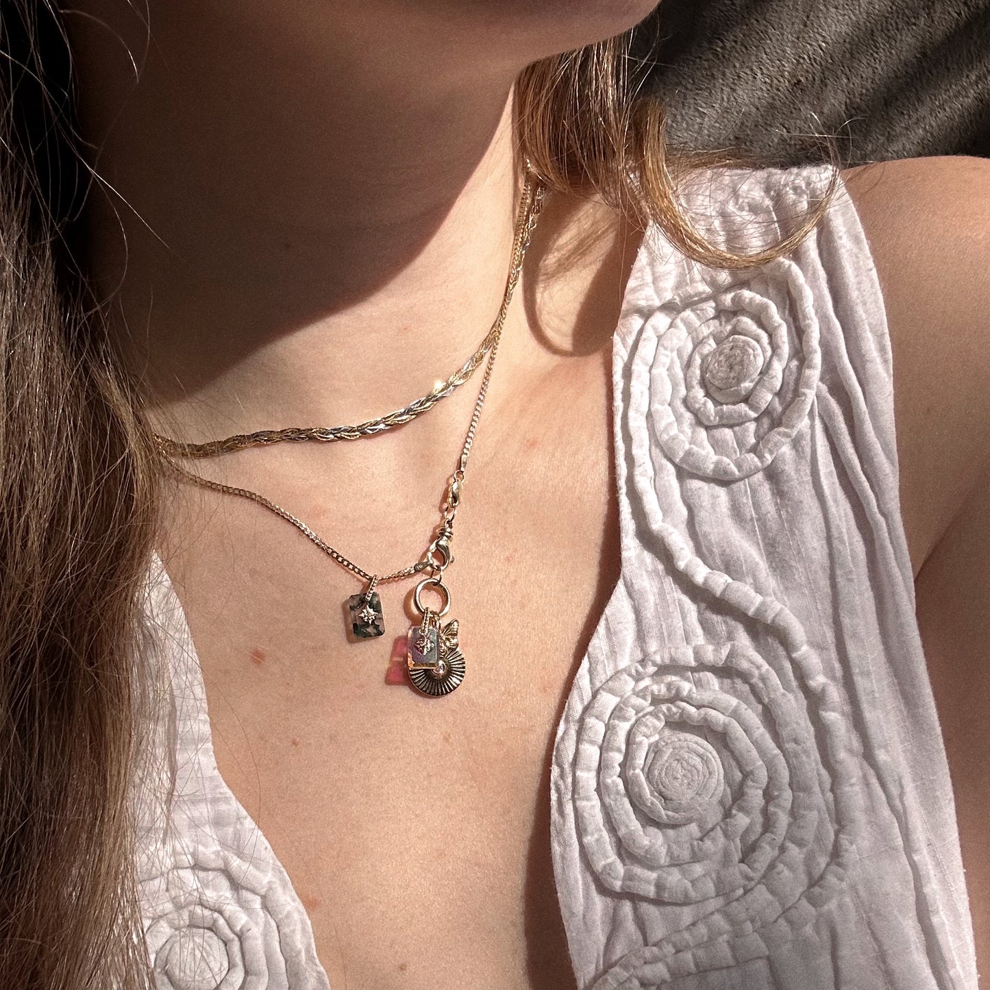 A woman in a white textured sleeveless top wears two gold necklaces—one is the Solid Gold Crystal Amulet Charm for Manifestation with a small square pendant—while basking in natural sunlight.