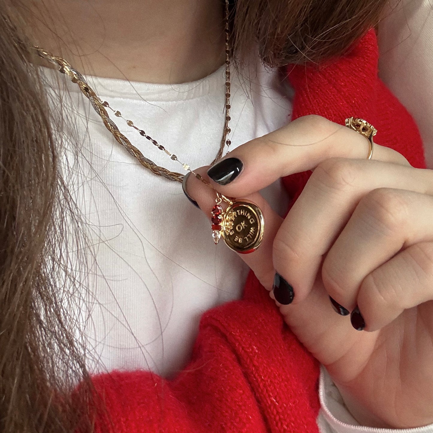 A person with black nail polish holds the Solid Gold Modern Ombre Birthstone Charm on a chain necklace, wearing a white top, red cardigan, and gold snake ring. The charm features engraved text and a small red gemstone birthstone.