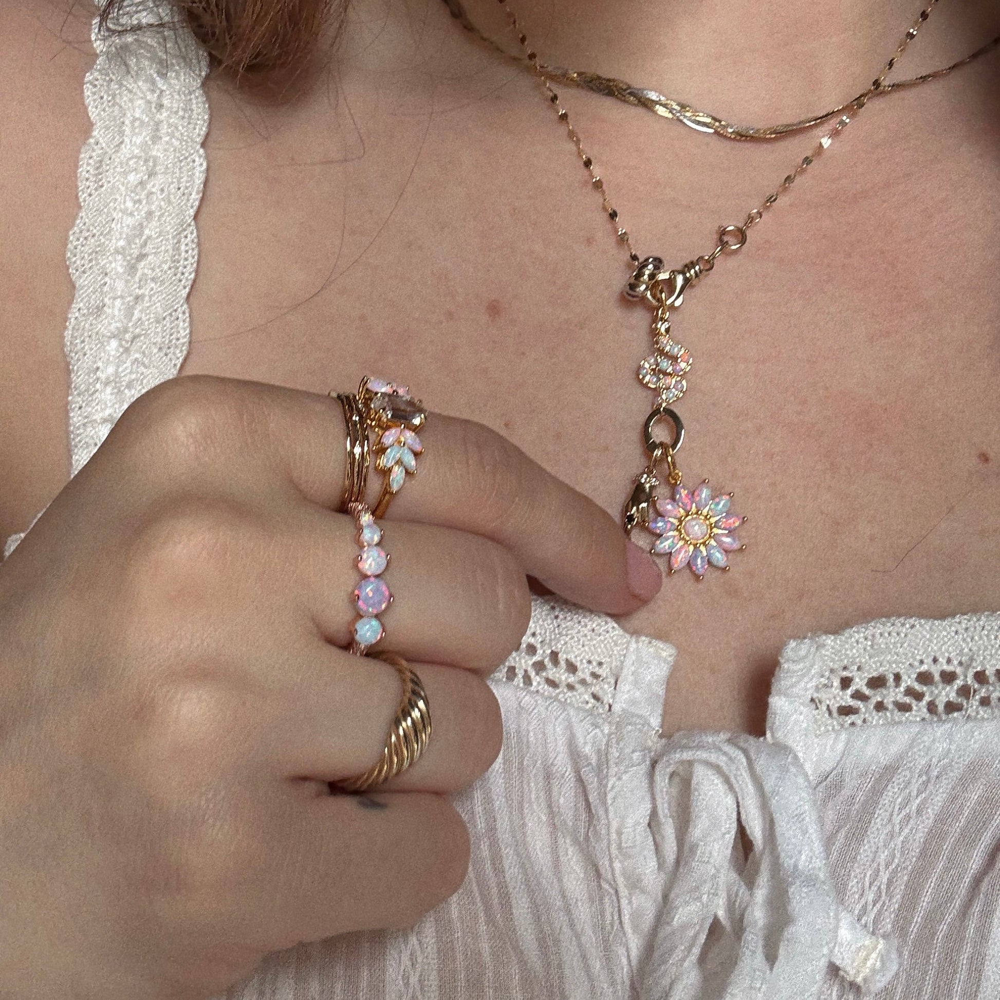 A person in a white lace top showcases Charms by La Kaiser gold jewelry, featuring 18k gold vermeil layered necklaces with a pink and white flower pendant, and gold rings with pastel pink and white stones.
