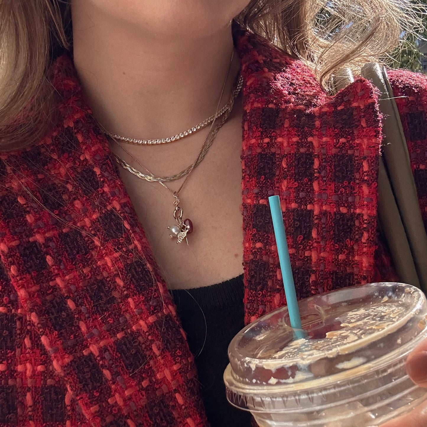 Wearing a red textured jacket and layered gold necklaces, including the Solid Gold Celebration Charm with a heart pendant, someone holds an iced drink with a blue straw as sunlight highlights their hair and clothing.