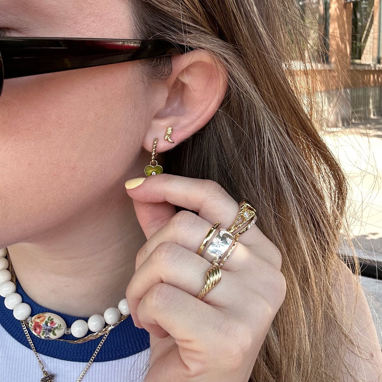 A person wearing black sunglasses, gold rings, a pearl necklace with a floral pendant, and 14k gold plated Cowboy Boot Studs holds their hand near their ear.
