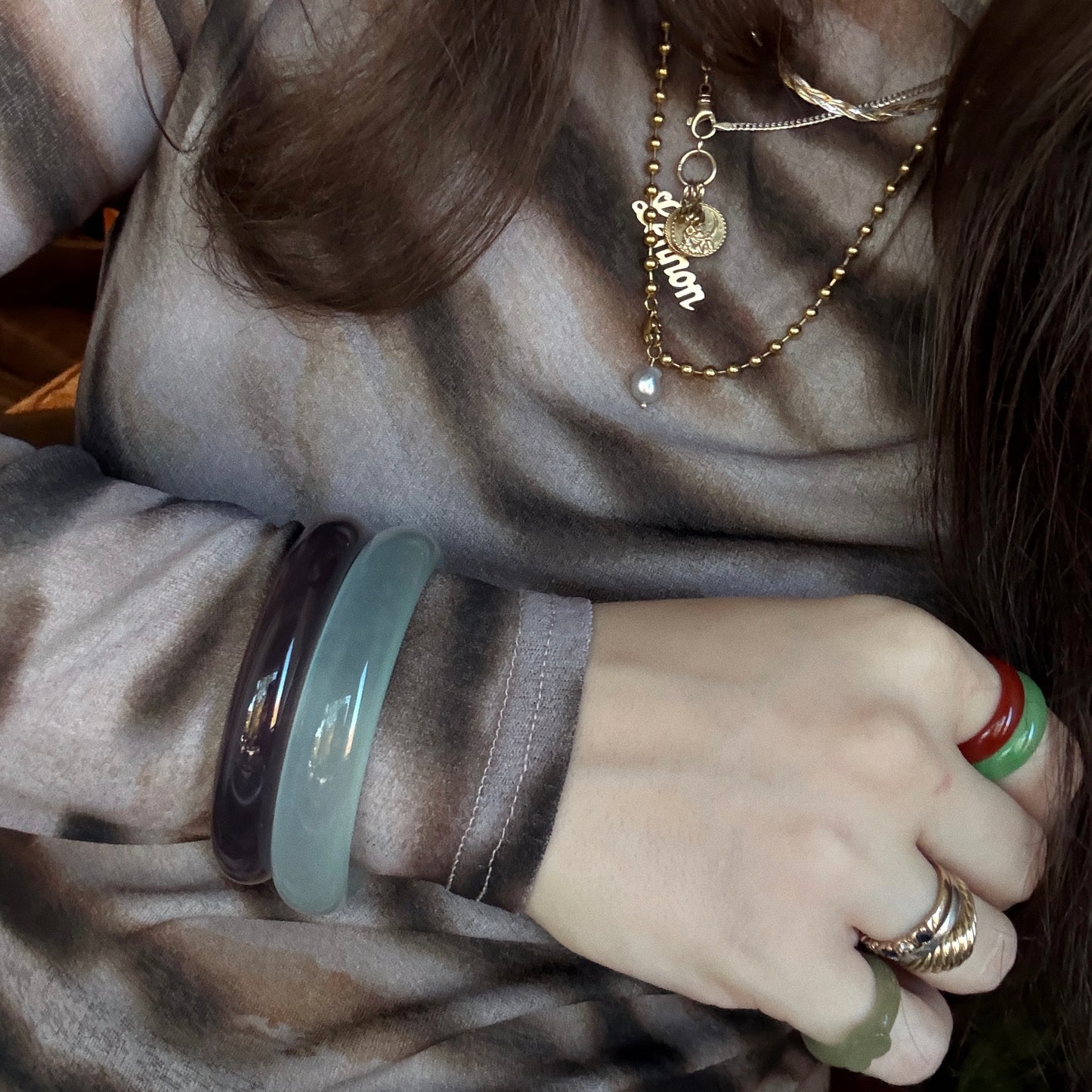 A person wears a sheer patterned top, layered gold necklaces—one with a Lemon name pendant—jade bangles, and the Heart Jade Ring featuring natural nephrite jade with green and red accents.