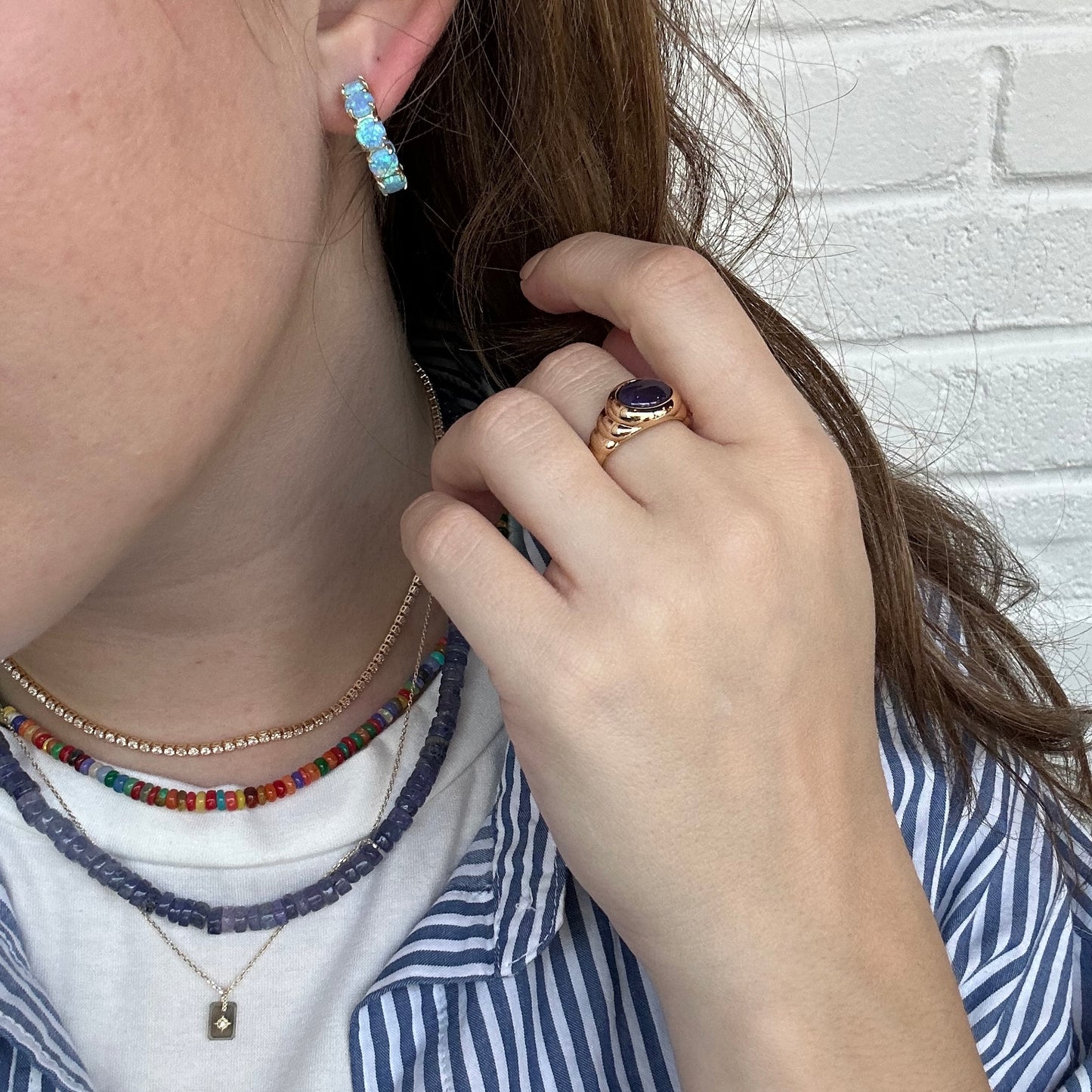 A person touches their hair, wearing three layered necklaces, an 18k yellow gold ring with a dark stone, and the Oh She Fancy Blue Opal Huggie Hoop Earrings. They have on a striped shirt over a white top against a white brick wall.