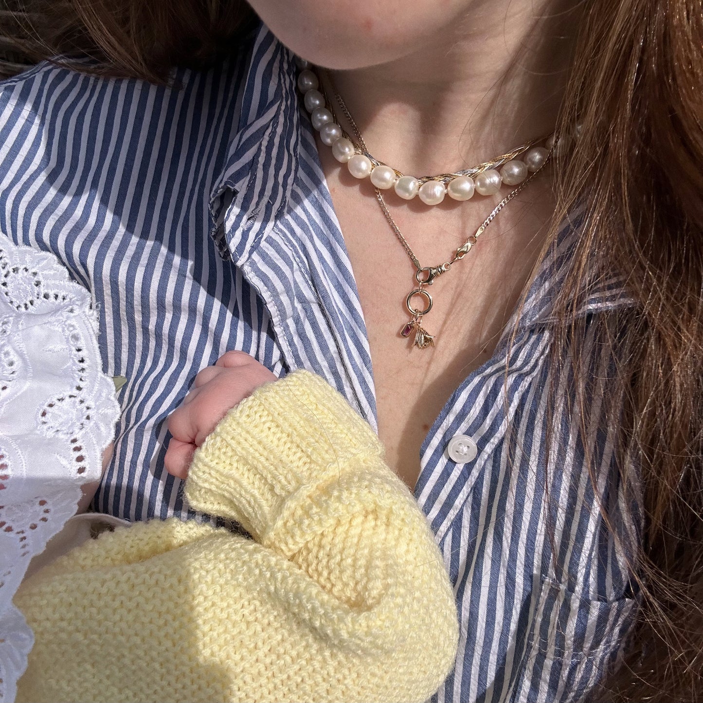 A woman in a blue and white striped shirt wears layered pearl and gold necklaces, featuring the Solid Gold Birth Flower Bud Charm, as a baby in a yellow knitted sweater rests against her chest, gently gripping her shirt.