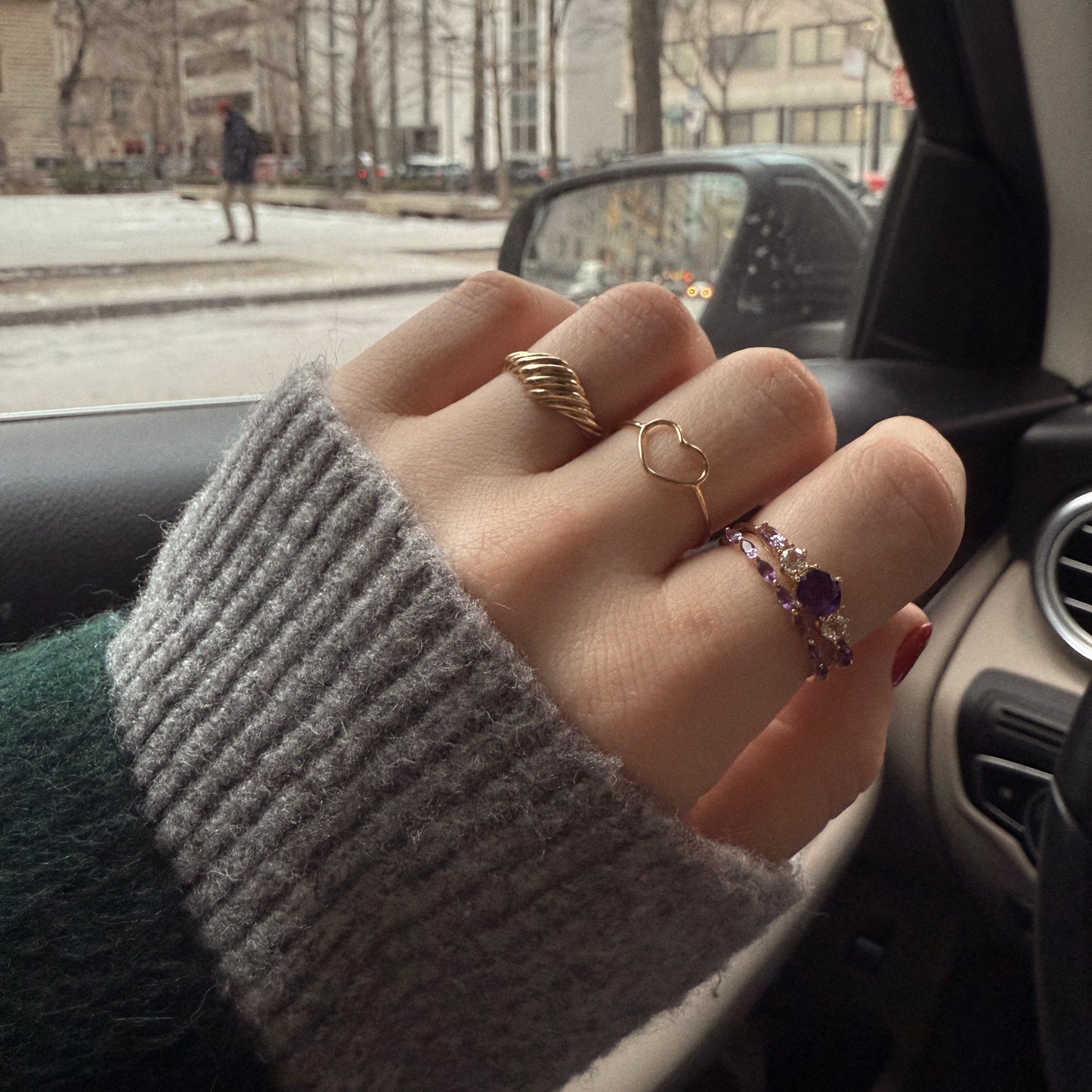 A close-up of a hand wearing several rings, including the Solid Gold Delicate Heart Ring, rests on a car door. The person’s gray knit sleeve and a blurred city street background add to the Family Gold charm.