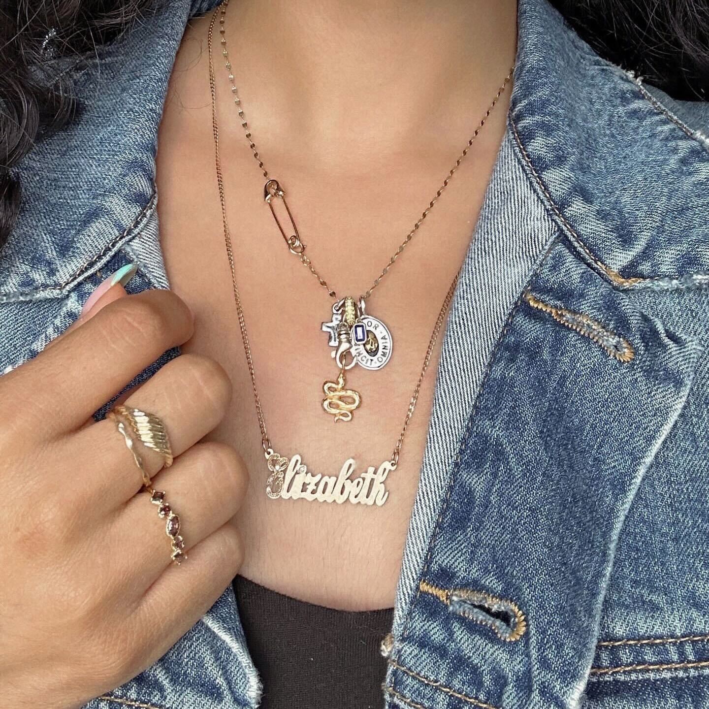 A person wears layered gold necklaces—one with Elizabeth, a snake charm, and vintage-inspired pieces—along with several rings, including the Solid Gold Twisted Signet Ring, and a denim jacket.