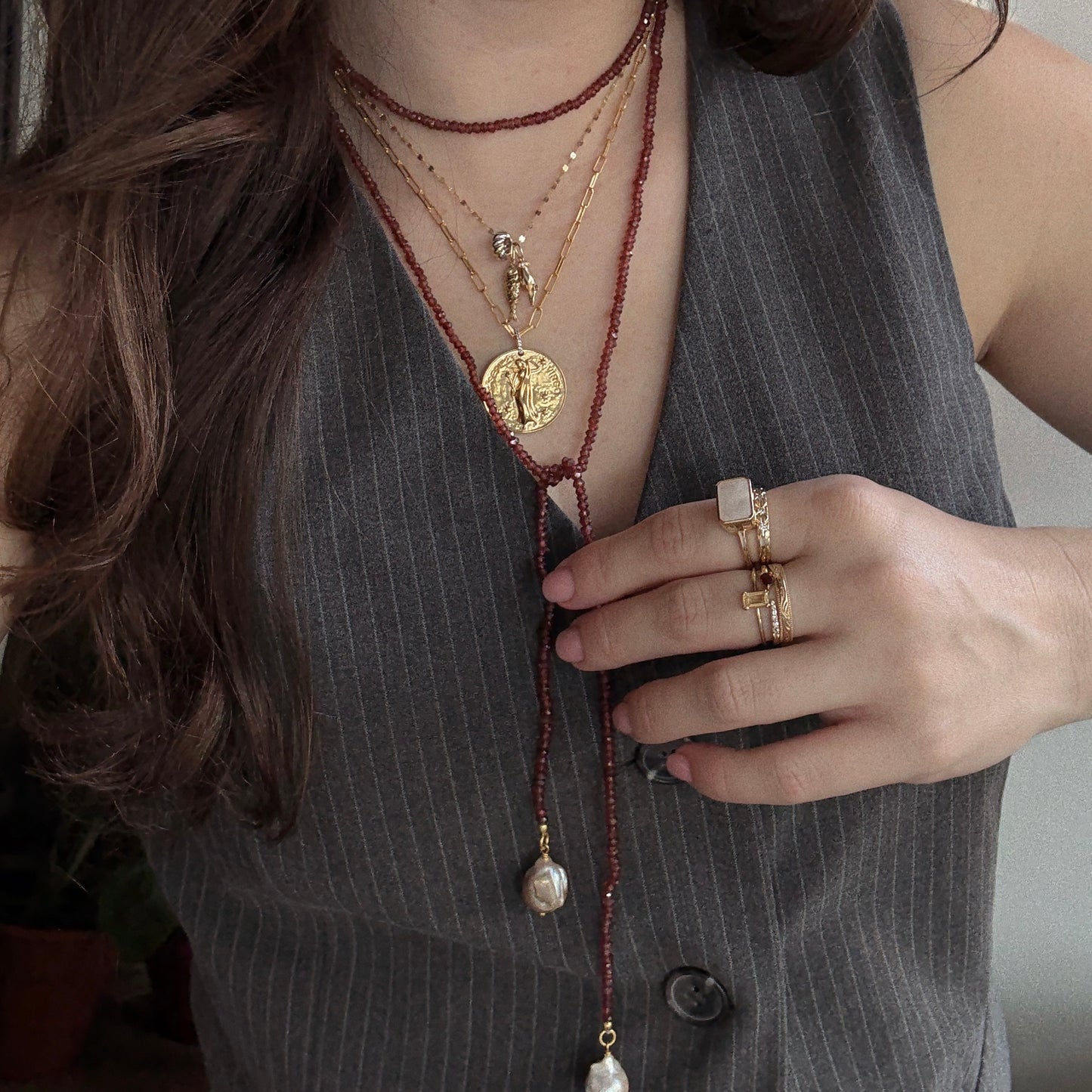 A person in a pinstripe vest layers gold necklaces, including the Gemstone Tie Necklace and a beaded pearl strand, with multiple gemstone gold rings and wavy hair draped over one shoulder.