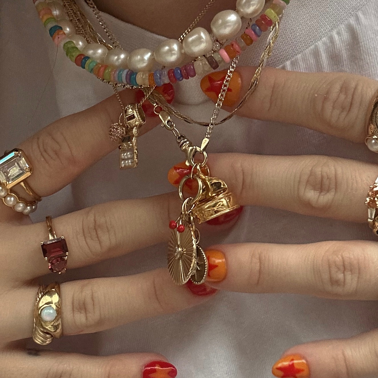 Close-up of hands with orange and red nail art, wearing gold rings and colorful layered necklaces featuring pearls, beads, and the standout Solid Gold Foodie Charm, all set against a white fabric background.