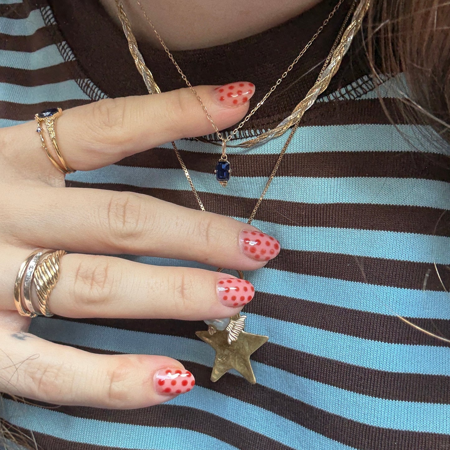 A hand with red and pink polka dot nails holds a solid gold necklace. Multiple gold rings and layered necklaces, including the Solid Gold 2025 September Capsule Classic Blue Sapphire Pendant Necklace, are worn over a brown and blue striped shirt.