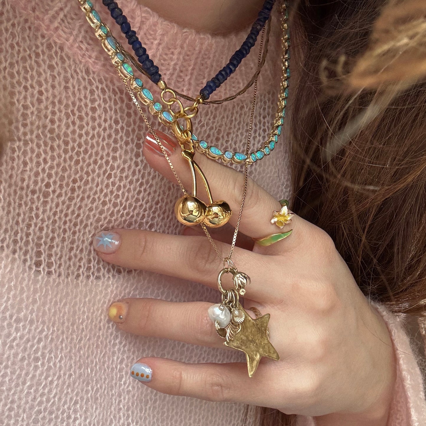 A person wearing Charms by Spicy Dan layered gold-plated necklaces, featuring a cherry charm, holds them with pastel nail art and a yellow flower ring. Paired with a light pink sweater—its a true Local Eclectic exclusive style.