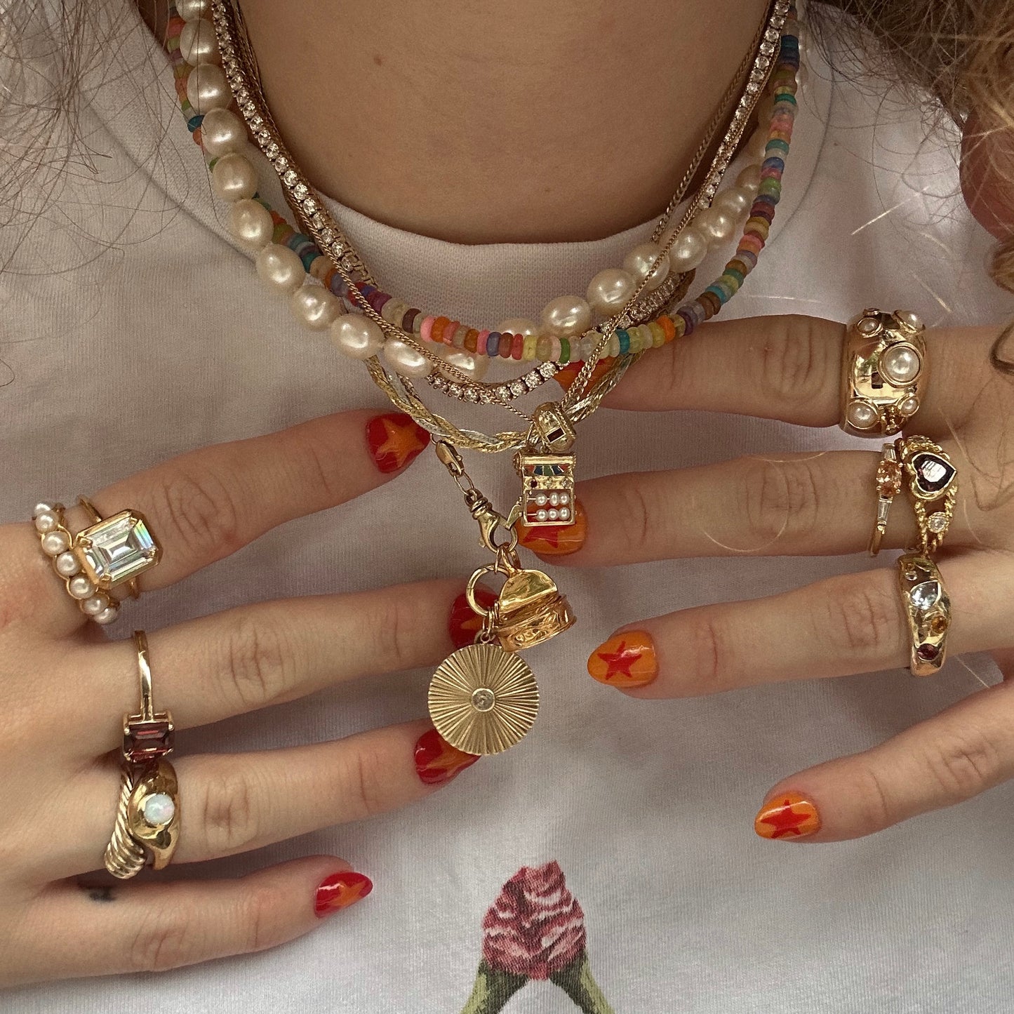 A person wears Charms by Hunt of Hounds layered with gold and pearl rings, colorful star nail polish, and a white floral print shirt. The necklaces feature pearls, beads, gold vermeil pendants, and playful charms.