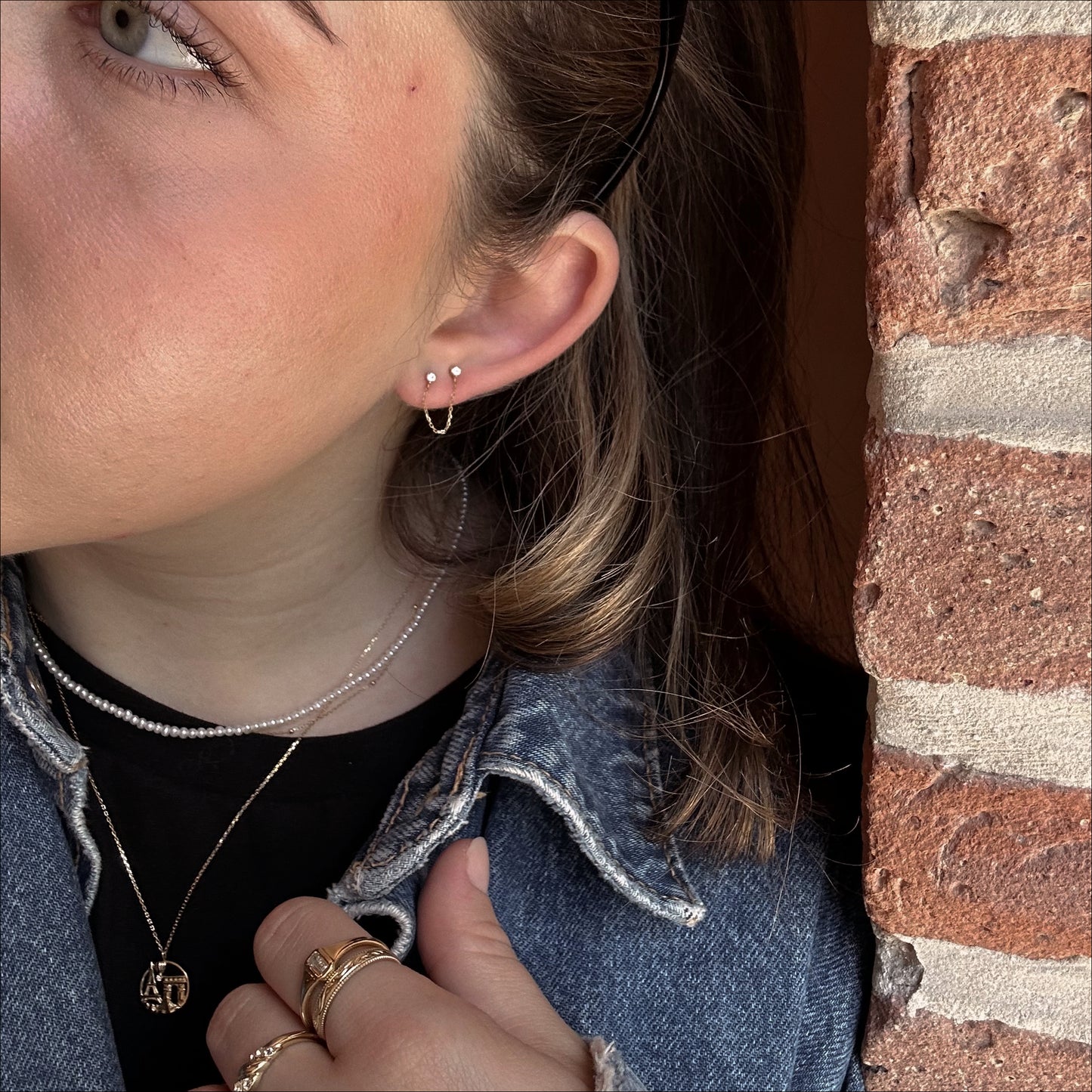 A close-up of a person wearing layered necklaces, gold rings, and a denim jacket, showcasing Solid Gold Double Piercing Chain Earrings against a brick wall backdrop.