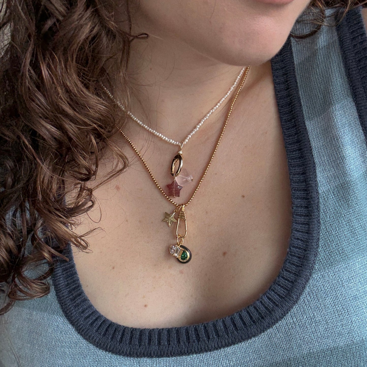 A close-up of a person with curly brown hair wearing a blue and gray striped top and three layered necklaces, featuring the Vermeil Beaded Chain along with beads, a pink crystal, a star charm, and a green gem pendant.