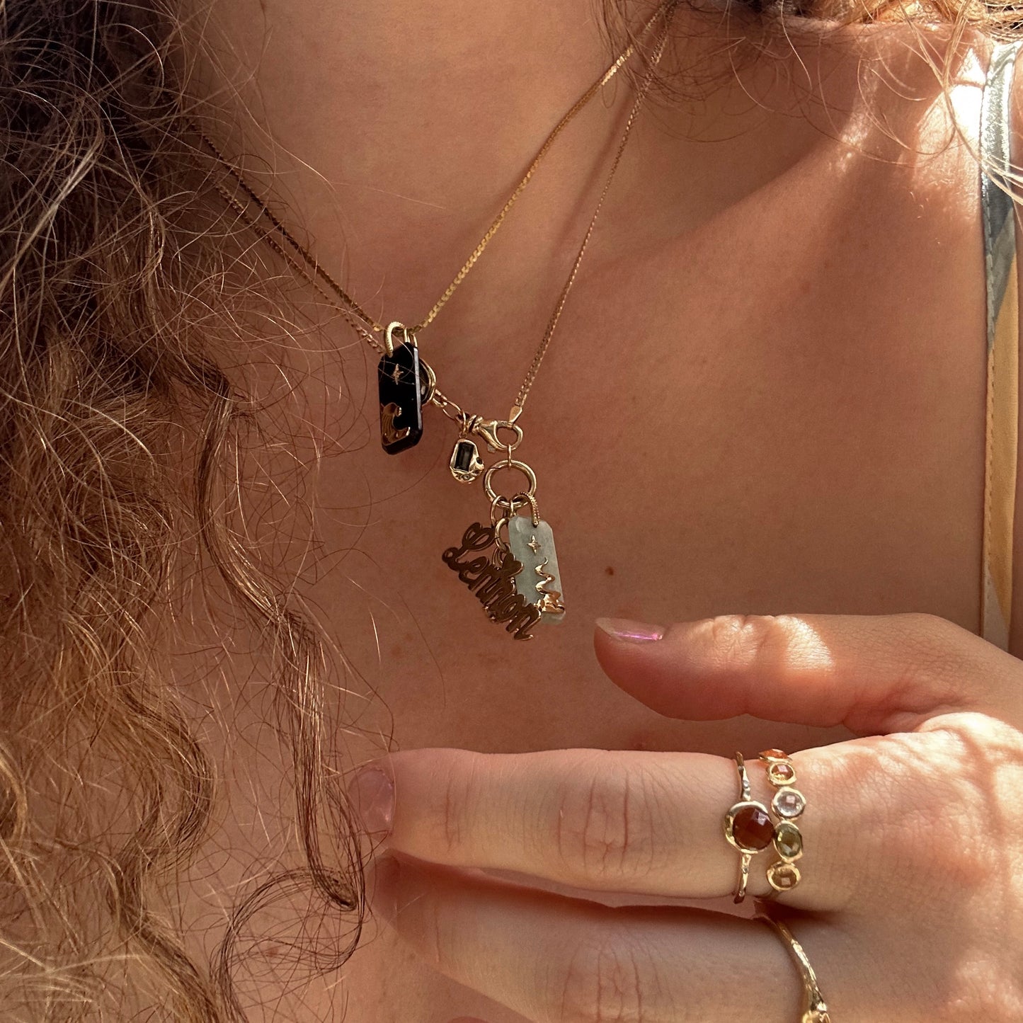 A close-up of a person’s neck displays layered gold necklaces, featuring the Solid Gold Amulet Charm from Local Eclectic and a second necklace with a dark stone. Their hand, adorned with gemstone rings, gently holds the gemstone jewelry.