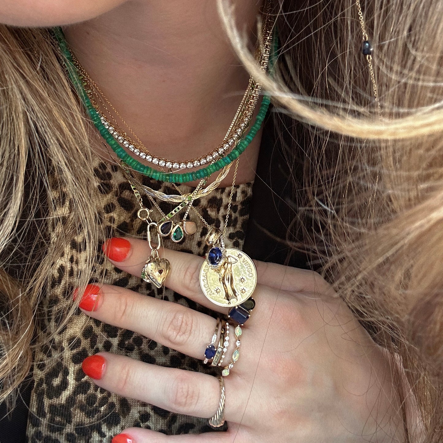 A person with wavy blond hair wears layered gold necklaces, including the Vintage Inspired Zodiac Coin Necklace, several gold rings with blue stones, and red nail polish. Their outfit features a leopard print pattern.
