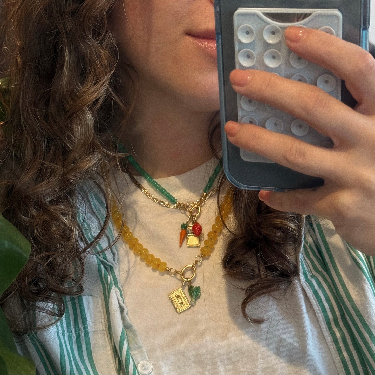 A person with wavy brown hair takes a mirror selfie wearing the Beaded Gemstone Charm Holder Necklace with carrot, book, and fruit charms. They have on a white shirt, green striped overshirt, and hold a phone with a blue pop-it case.