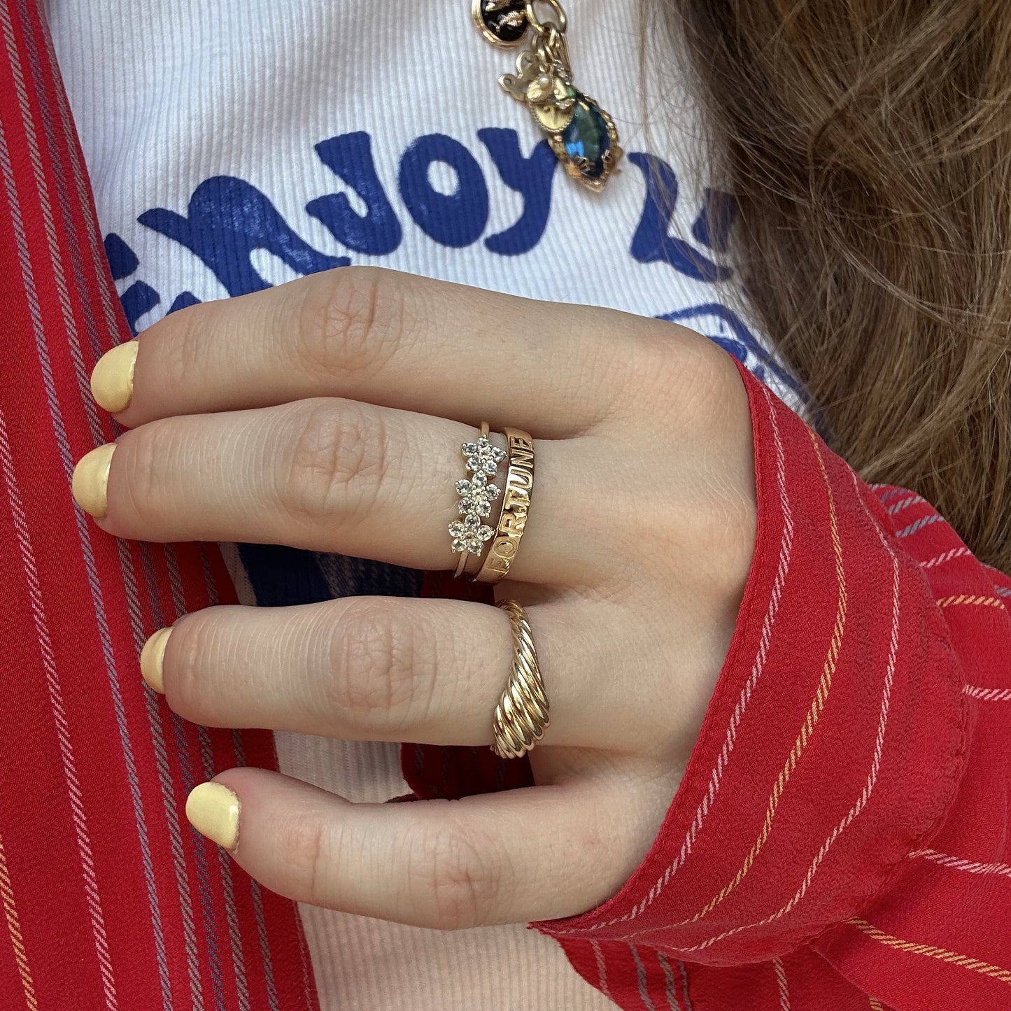 A hand with yellow-painted nails wears three gold rings, including a Solid Gold Blue Topaz Flower Trio Ring and one engraved with LOVE. The person is dressed in a red striped shirt, white T-shirt with blue lettering, and a Family Gold charm necklace.