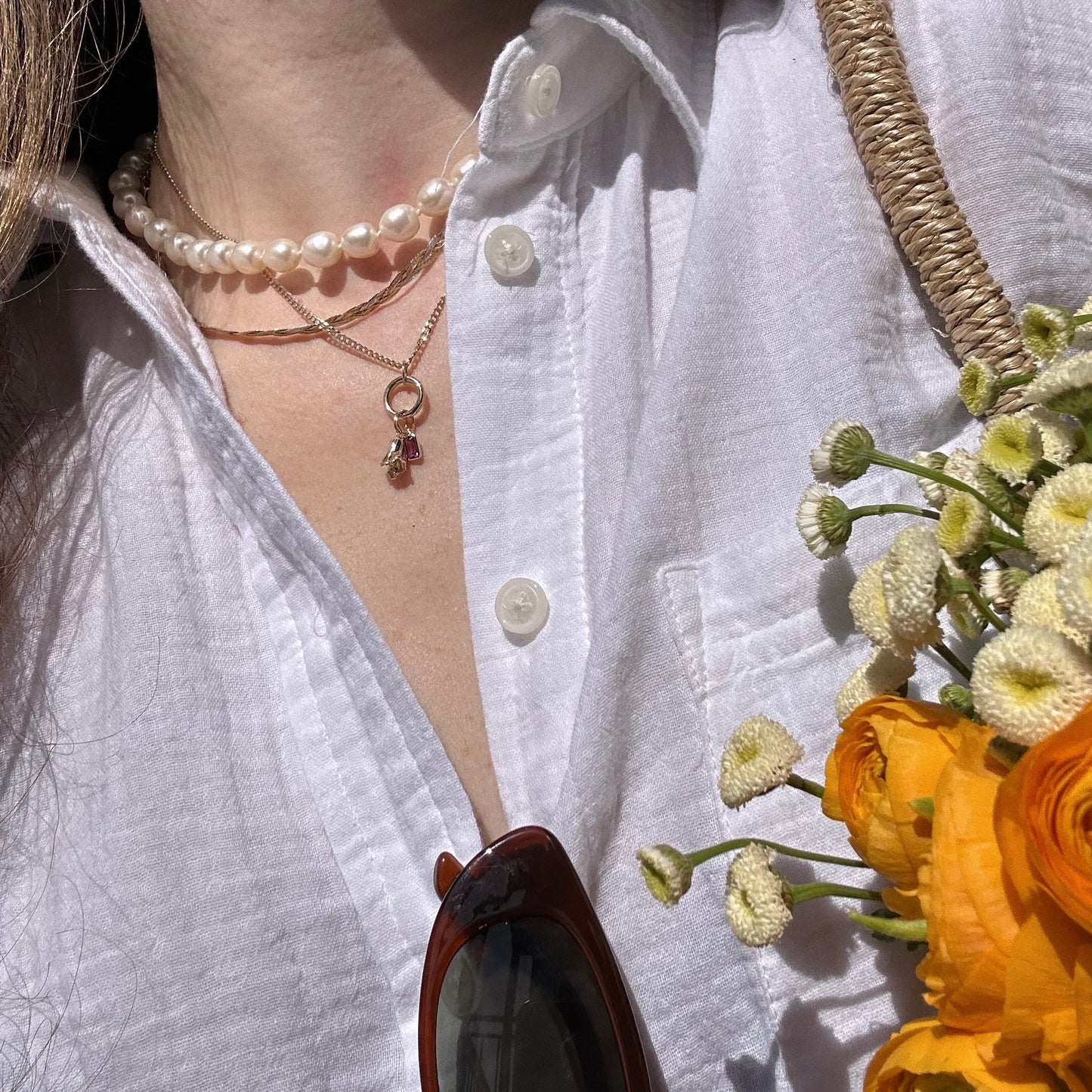 Close-up of a person in a white shirt, layered pearl and gold necklaces, and carrying yellow and white flowers. A Solid Gold Birth Flower Bud Charm adds personal style. Brown sunglasses hang from the shirt; a woven bag rests on the shoulder.