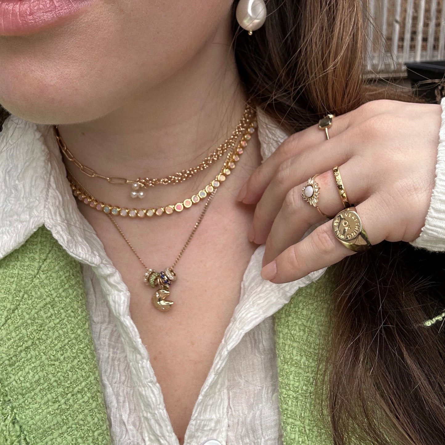 A woman wears layered gold necklaces, pearl earrings, and several gold rings featuring the Solid Gold Anniversaire Charm, styled with a textured white shirt and a light green knit vest.