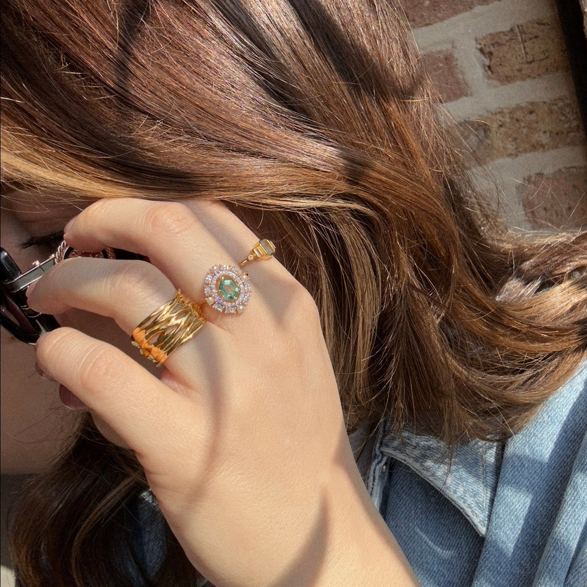 A person with wavy brown hair adjusts black sunglasses, wearing a 14k gold vermeil ring and a Fern Green Topaz Statement Ring with Pearl Halo, paired with a denim jacket against a brick wall backdrop.