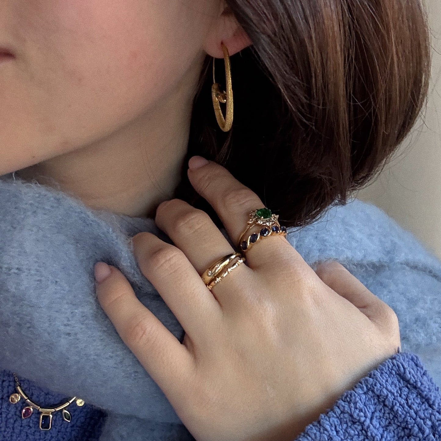 A woman with short brown hair wears Mini Snake Hoops, several gold rings with colorful gems, and a blue scarf. She touches her neck, and her sweater is adorned with a decorative gemstone brooch.