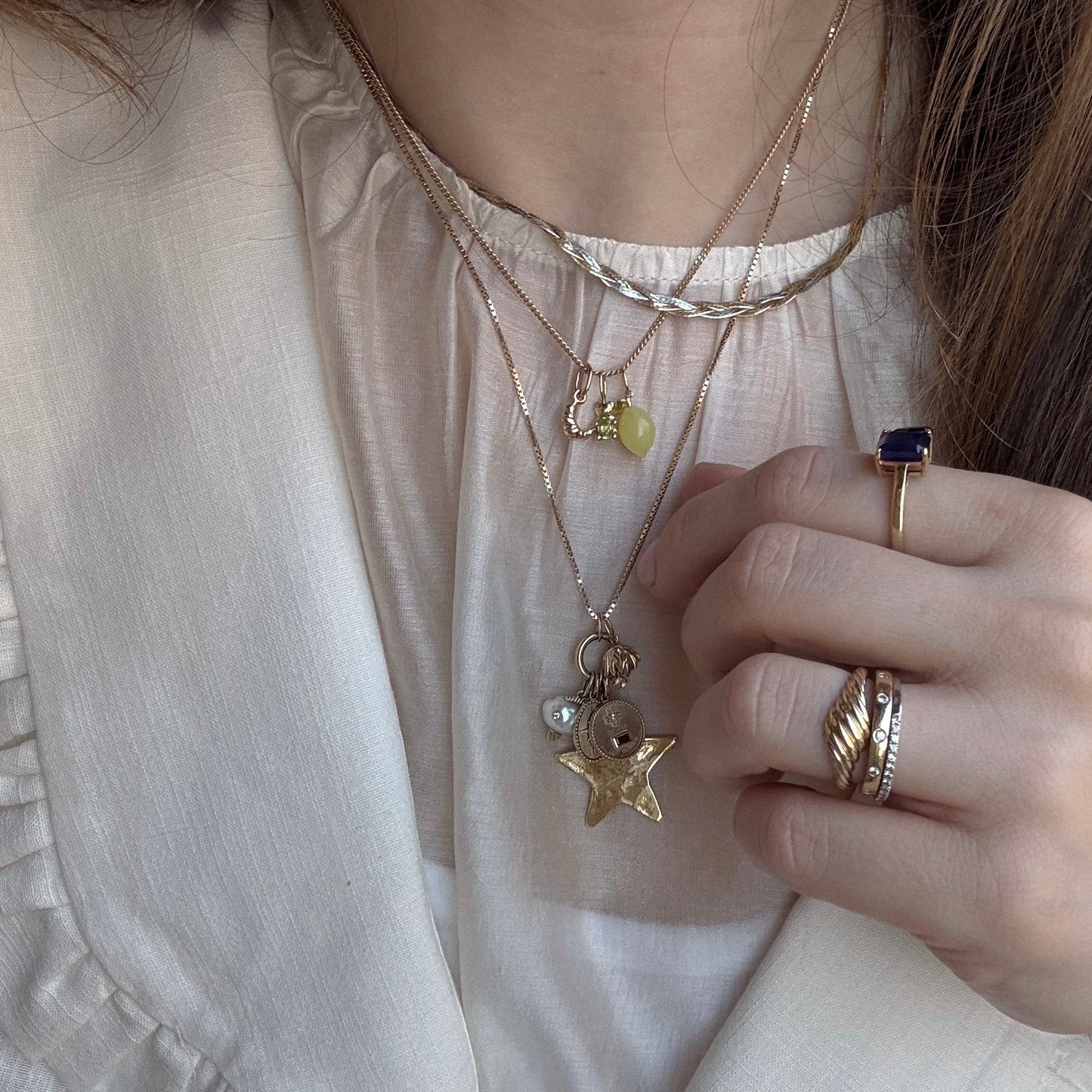 A person wearing layered 9k yellow gold necklaces with various pendants, including the Solid Gold Foodie Charm, a star, a pearl, and a yellow stone, plus gold and blue rings, dressed in a cream-colored blouse.