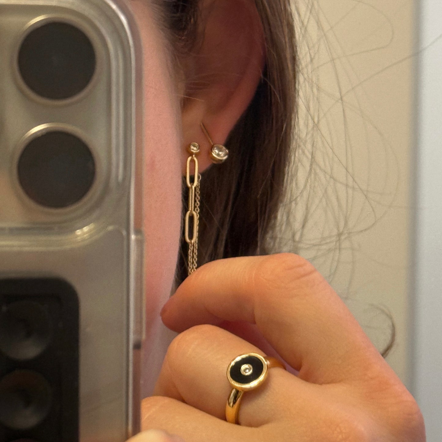 A person takes a mirror selfie, showing two earrings and holding their earlobe with the Balia Black Enamel Ring. Part of their phone appears in the foreground.