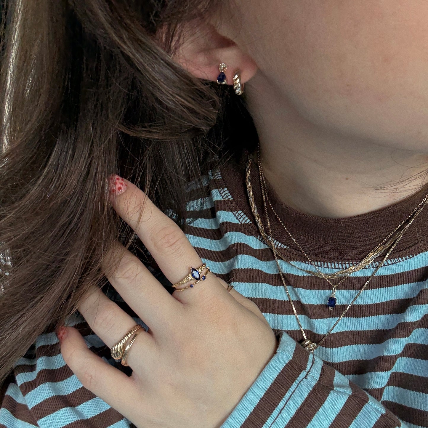 A person wears the Solid Gold 2025 September Capsule Blue Sapphire Statement Set, featuring layered necklaces and rings with sapphire stones, paired with a striped blue-brown shirt and small gold earrings. They have wavy brown hair and pink nails with red spots.