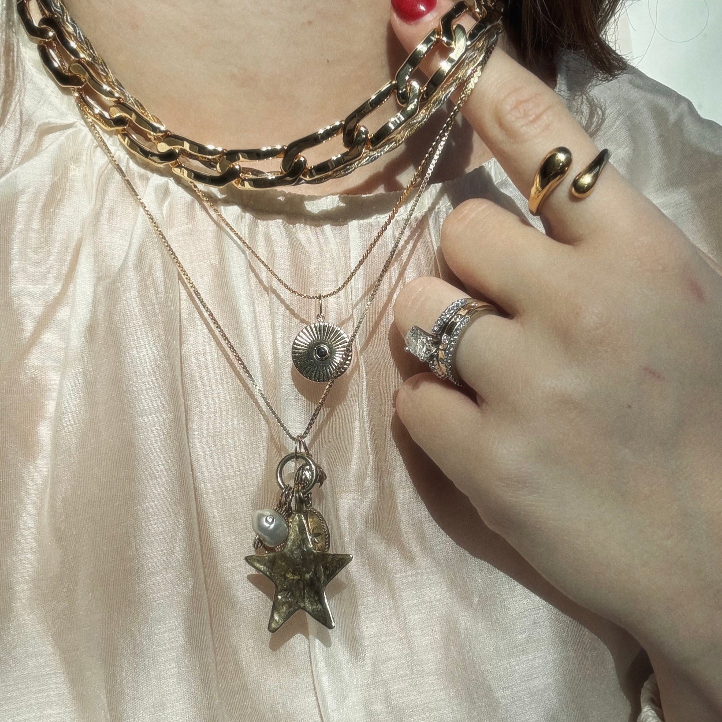 A person wearing layered 14k gold plated and silver necklaces, including a star pendant and the Mantra Empowerment Coin Charm, plus multiple rings on their fingers, poses against a cream-colored satin blouse.