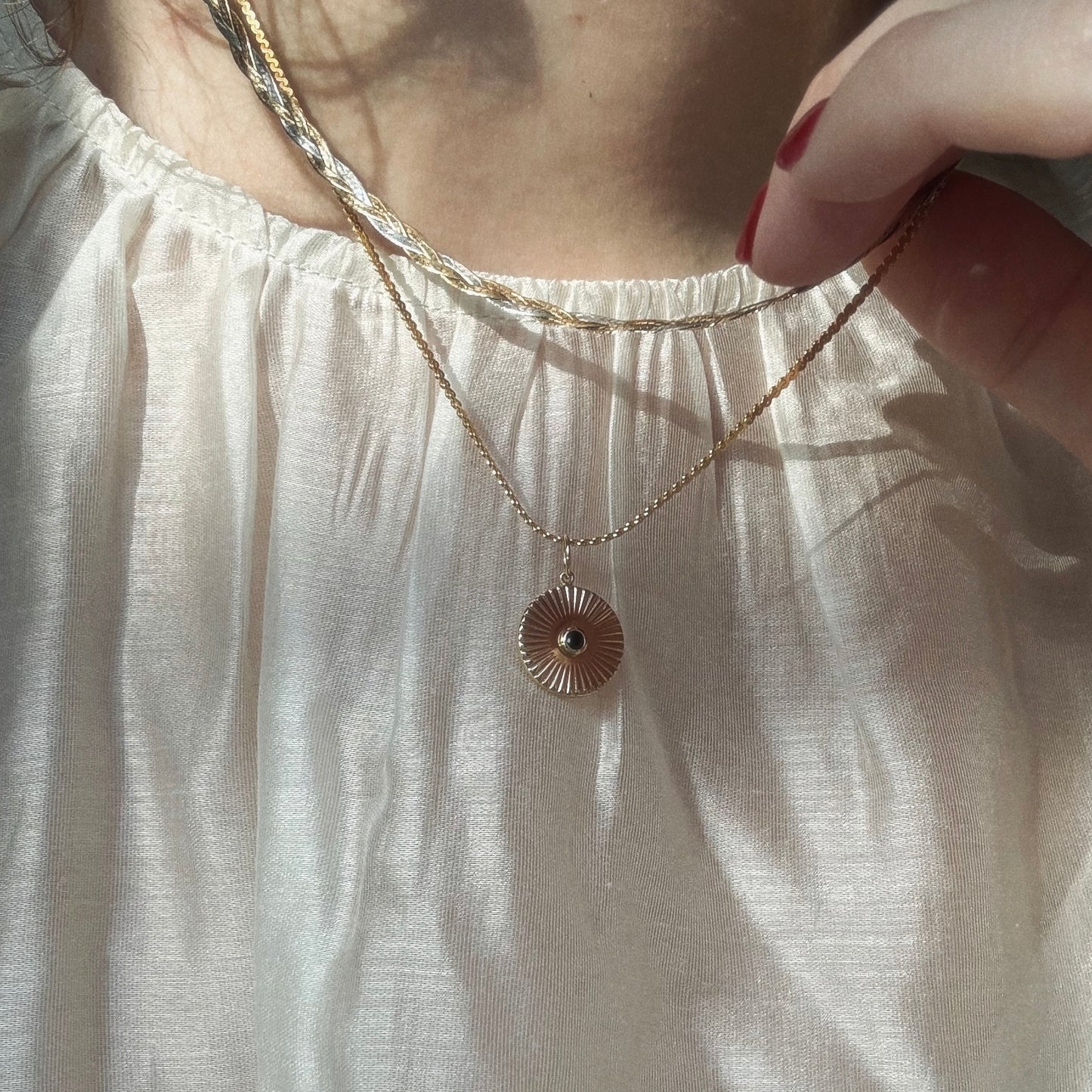 A person in a cream, slightly sheer blouse holds the Mantra Empowerment Coin Charm necklace in 14k gold plating. Sunlight highlights the mantra jewelry and fabric, with only their lower face and neck visible.