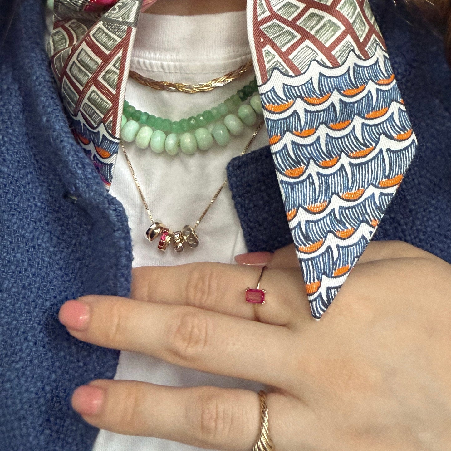 A close-up of a person wearing a blue textured jacket and colorful collar, showcasing layered necklaces with the Solid Gold Anniversaire Charm, rings, and manicured light pink nails as their hand rests on their chest.