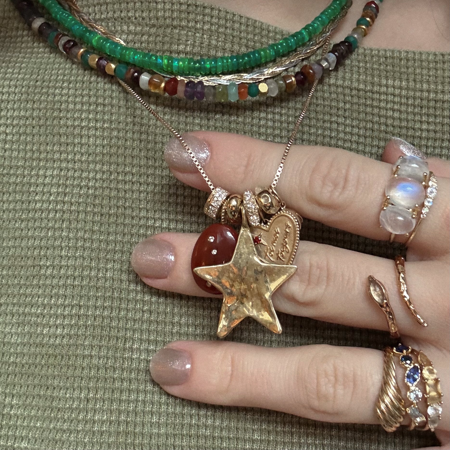 A hand with metallic nail polish holds layered necklaces with colorful beads, a gold star pendant, Solid Gold Spacer Charms, and yellow gold charms. The person wears multiple gemstone rings and a textured green top.